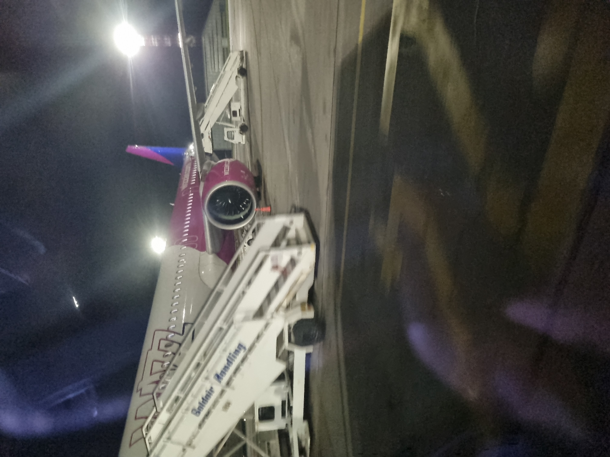 The image shows an airplane parked at an airport, likely at night, based on the ambient lighting and dark sky visible. The airplane is white with pink and blue accents on the tail. Stairs are positioned next to the airplane, indicating that passengers are boarding or disembarking. The stairs bear the name "Safdair Handling." The setting appears to be an airport apron, with visible tarmac and possibly other airport infrastructure nearby. The location is identified as the city of Sofia, Bulgaria, but the exact location within the airport is not specified. No people are visible in the image.