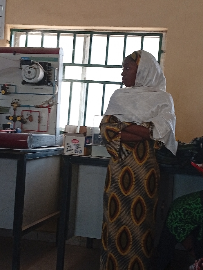 A woman in a patterned brown and gold dress with a white head covering stands in a room with industrial equipment. She has her arms crossed and is looking to the right. Behind her is a large window with multiple panes. To the left is a piece of industrial machinery mounted on a cabinet, and next to it are some boxes and equipment on a counter. Some fabric is visible on the right.