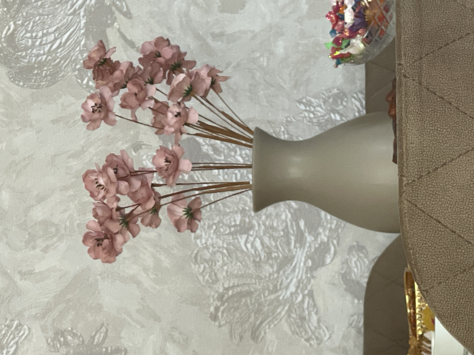 A bouquet of artificial pink flowers in a beige vase sits on a table in front of a textured wallpaper. To the right of the vase, a glass bowl filled with colorful candies is visible. In the foreground, a portion of a quilted brown surface is present. The image is a close-up shot, and no specific location in Sumgayit, Azerbaijan can be determined.