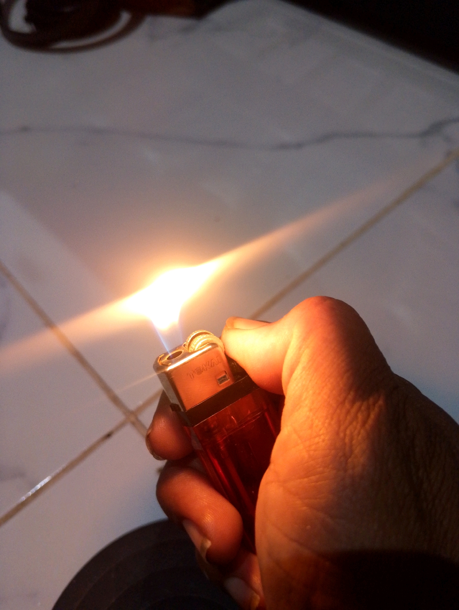 A hand in Sumbergirang, Indonesia, is holding a red lighter that is actively burning, producing a bright flame and light. The lighter has "TOKAI" printed on its metallic top. The background consists of a tiled floor, with some of the tiles appearing to have faint cracks or lines. The lighting suggests it is either dusk or the image is taken in a dimly lit indoor space with a light source creating a strong glare across the scene. The image is framed from a close-up perspective, focusing on the hand and the lighter.