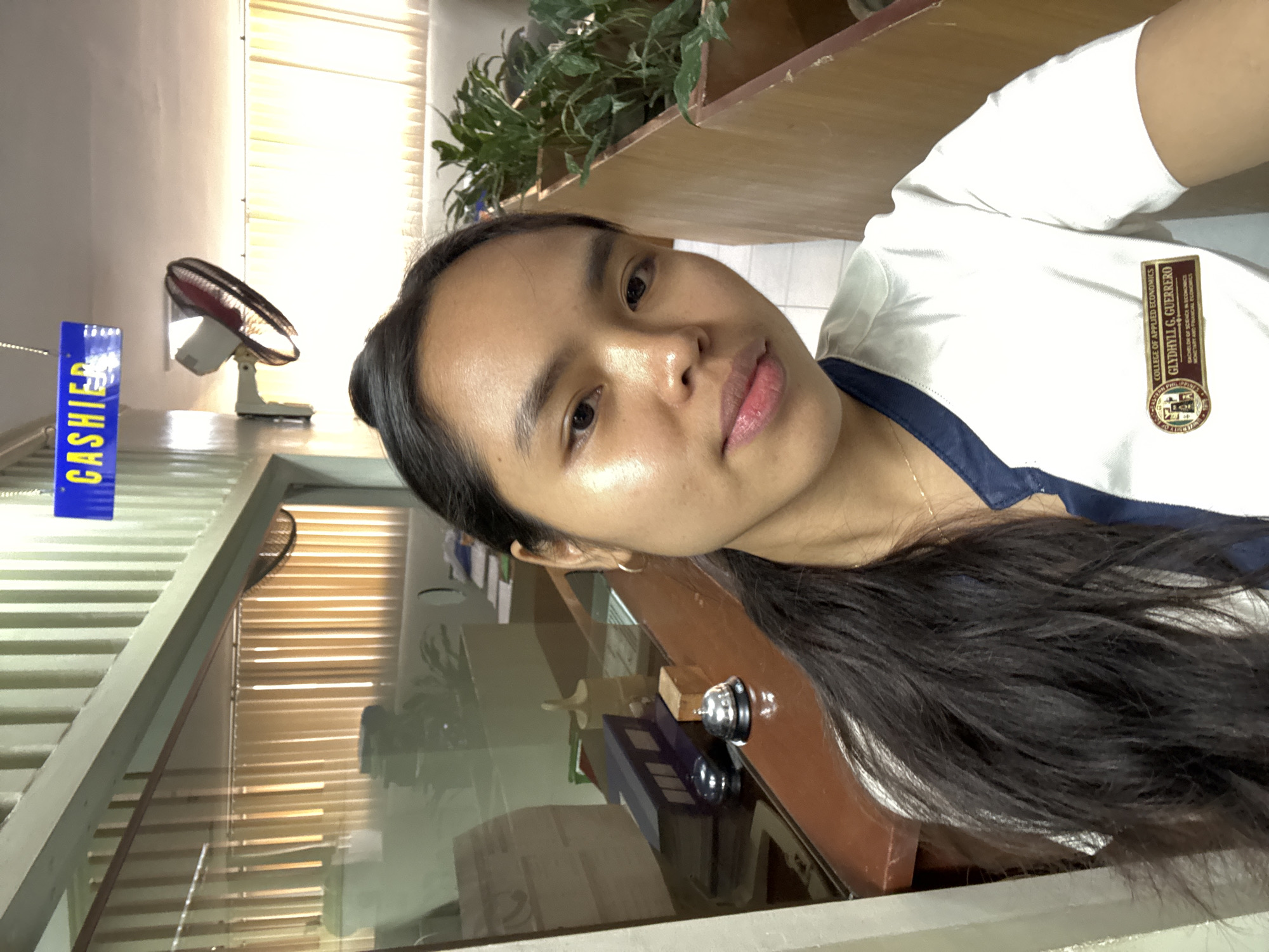A young woman with dark hair pulled back is posing for a selfie. She is wearing a white uniform with a blue collar and a name tag that reads "COLLEGE OF APPLIED ECONOMICS GLYDHYLL G. GUERRERO BACHELOR OF SCIENCE IN ECONOMICS MONETARY AND FINANCIAL ECONOMICS". She is standing in front of a counter with a glass partition. Behind her, on the counter, are two service bells and some folders. A sign reading "CASHIER" is visible overhead. The background shows a room with vertical blinds and potted plants, consistent with an office or administrative setting. The overall environment suggests a public institution or business. While the image is set in the city of Davao City, Philippines, no specific landmarks or context are provided to pinpoint an exact location within the city.