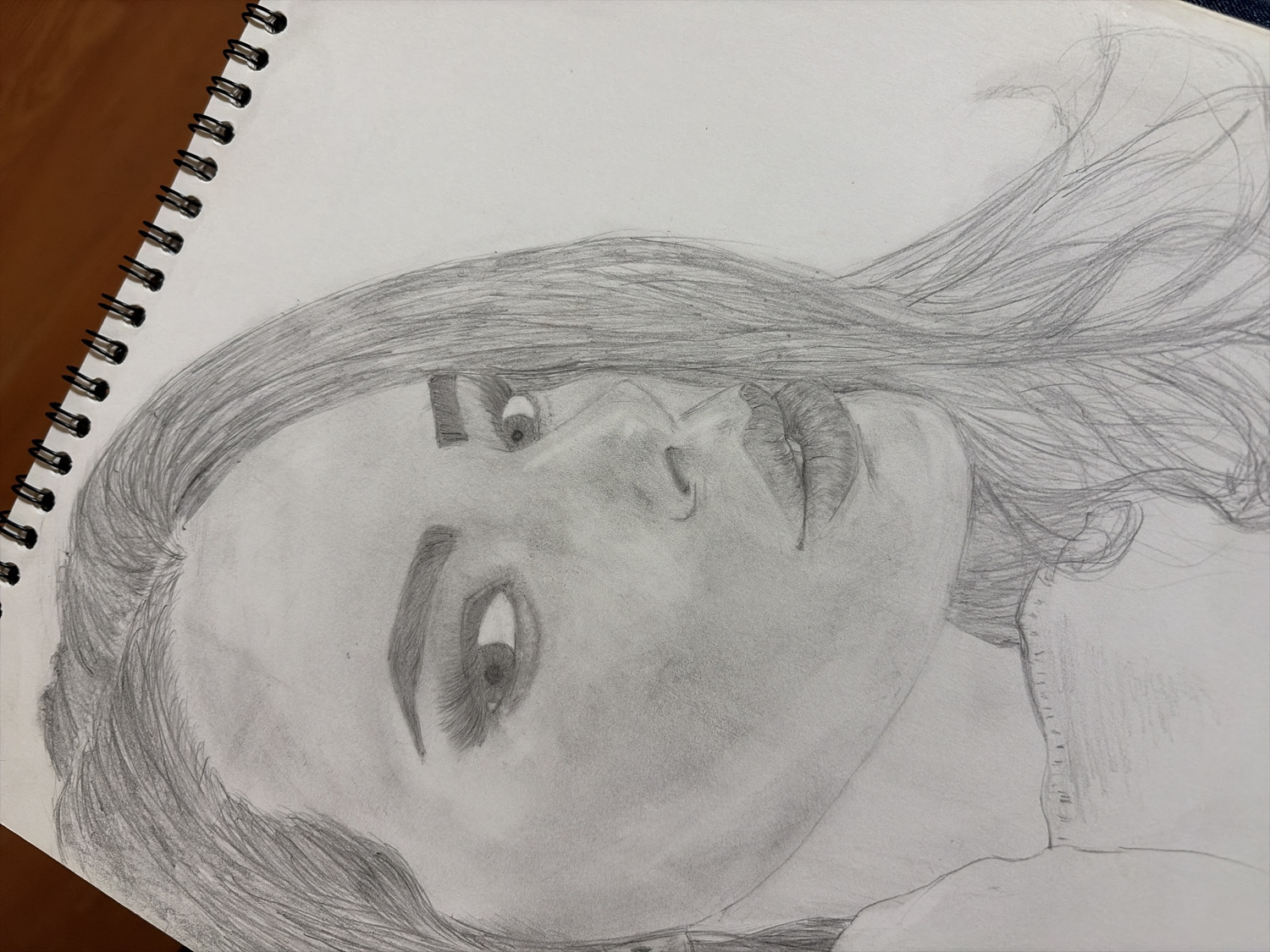 A realistic pencil drawing of a person, likely a woman, is depicted on a white page within a spiral-bound sketchbook. The drawing portrays the subject from the chest up, with her head tilted upwards and to the right from the viewer's perspective. Her long, dark hair flows around her face and over her shoulders, rendered with detailed individual pencil strokes that convey movement.
The subject's face shows well-defined features, including expressive eyebrows, open eyes with visible pupils and eyelashes gazing upwards and slightly to the left. Her nose is prominent, and her full lips are slightly parted, suggesting a pensive or serene expression. Varied shades of graphite are used effectively to create depth, contour, and highlights on the face, indicating a light source illuminating her from the left or above.
The sketchbook itself rests on a brown wooden surface, partially visible along the left edge of the image, next to the black spiral binding. There is no discernible activity taking place other than the static portrait. No visible text is present in the image.