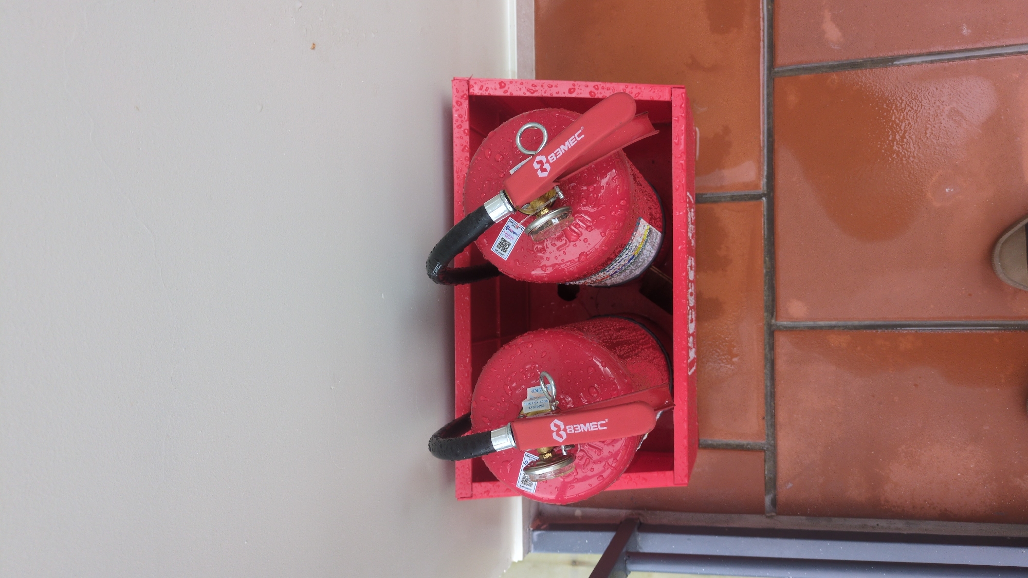 Two red fire extinguishers are positioned side-by-side in a red metal holder. Both fire extinguishers are cylindrical with black hoses and red handles. They are covered in water droplets. The holder is placed on a floor tiled with reddish-brown square tiles. Above the holder is a light-colored wall. To the left of the holder is a grey metal railing. The scene appears to be outdoors or in an area exposed to the elements, as indicated by the water droplets and the tiled flooring. There is no visible indication that this scene is located in Da Lat, Vietnam.