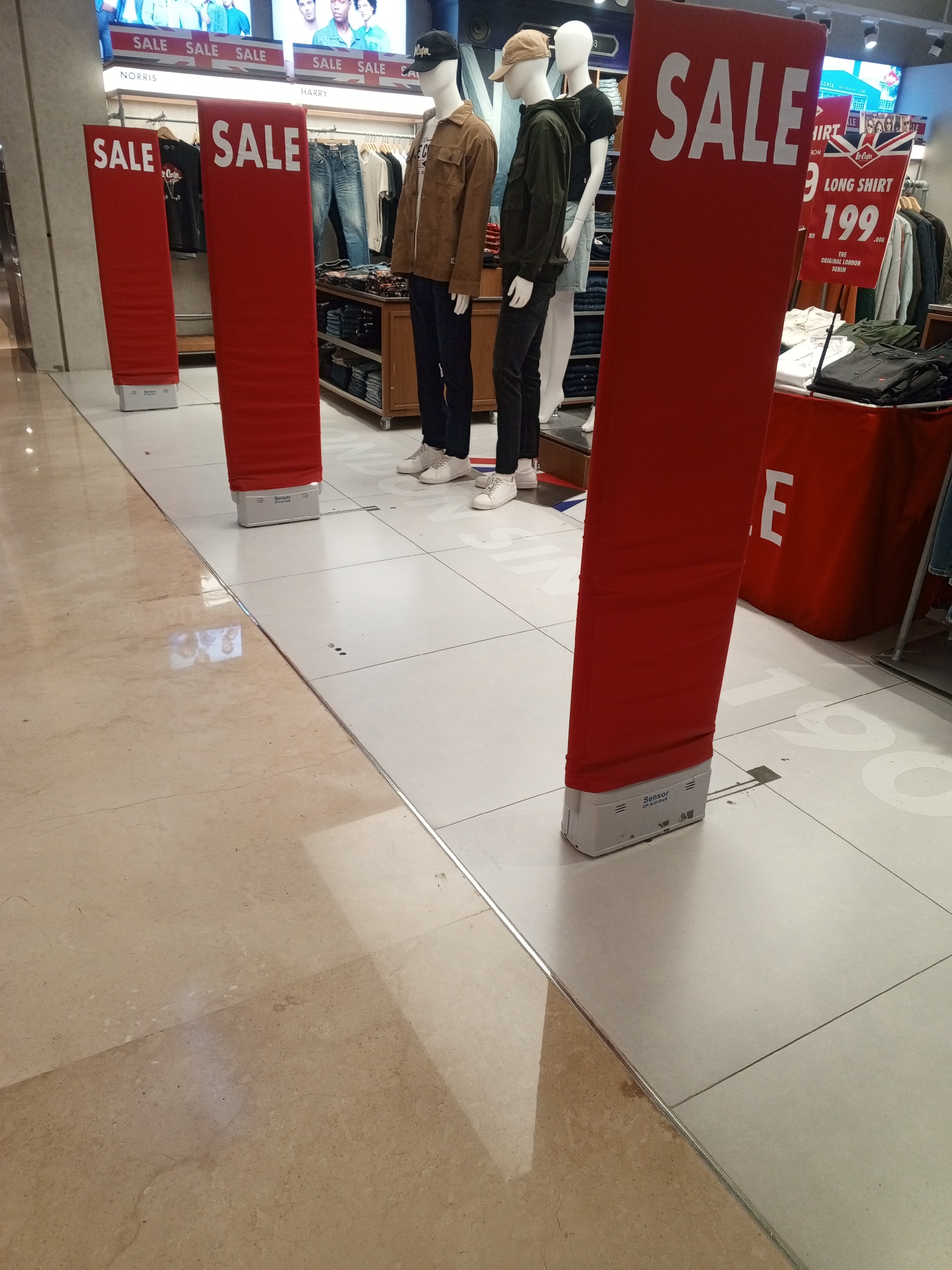 The image depicts the interior of a clothing store, likely a department store, with a prominent "SALE" display. In the foreground, four large, vertical red banners with white "SALE" lettering are positioned on the floor. The banners are rectangular and extend upwards, partially obscuring the merchandise behind them. The floor is tiled in a light color, with a polished beige marble-like section to the left.

In the background, several mannequins are dressed in casual clothing, suggesting men's apparel. One mannequin on the right wears a brown jacket, dark pants, and white sneakers. Another mannequin to its left wears a dark jacket and black pants, with a beige baseball cap. Behind the mannequins, racks of clothing are visible, including jeans and shirts. A wooden display shelf is also present, filled with folded clothing items.

Further in the background, a large advertisement board displays images of people and text, including "SALE SALE SALE". Another sign shows a product image and price information, with text reading "LONG SHIRT" and "199". The overall ambiance is that of a retail environment holding a promotional sale. The presence of sale banners and mannequins indicates a commercial setting focused on selling clothing.

This scene is located within the Cibinong City Mall in Cibinong, Indonesia, as indicated by a sign that says "Cibinong City Mall" in the upper left corner of the original image. The mall environment is suggested by the polished flooring and the arrangement of retail displays.