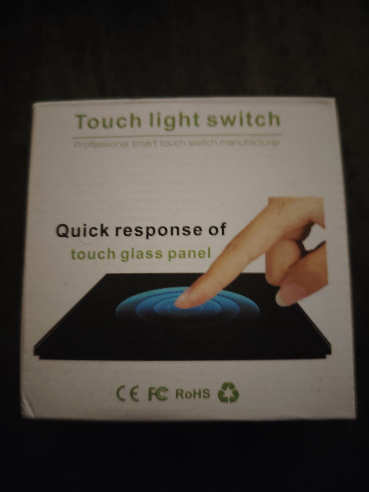 A white, rectangular cardboard product box is depicted from a slightly elevated, frontal perspective, resting on a dark, indistinct surface. The box displays product information and certifications. Prominently centered at the top, the text "Touch light switch" is printed in green capital letters, with "Professional smart touch switch manufacturer" in smaller, lighter green text directly below. The middle section features the phrases "Quick response of" in black, followed by "touch glass panel" in green. Below this text is a graphic illustrating a light-skinned index finger making contact with a black, rectangular touch panel, from which concentric blue light ripples emanate. The bottom of the box shows the green certifications "CE FC RoHS" and a green recycling symbol. The box exhibits minor scuffing and creasing along its edges, particularly the left side. The background is uniformly dark and out-of-focus, providing contrast for the brightly lit box. This scene is located in Huntingdonshire, United Kingdom.