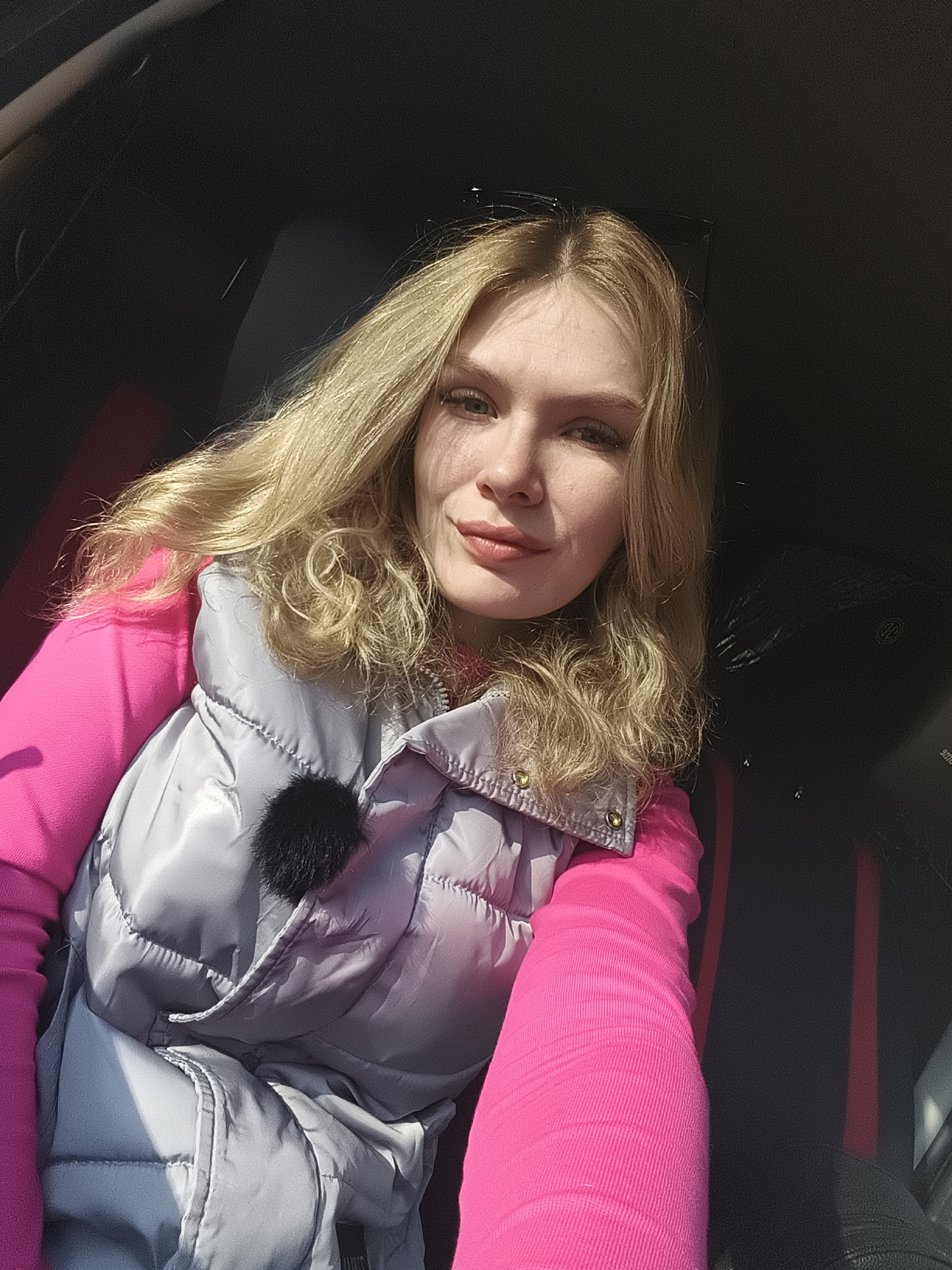 A woman with blonde hair is seated inside a car. She is wearing a pink long-sleeved shirt and a grey padded vest. The interior of the car is visible, showing dark upholstery with red stripes. The location is unknown, however the text included the city of Izhevsk, Russia.