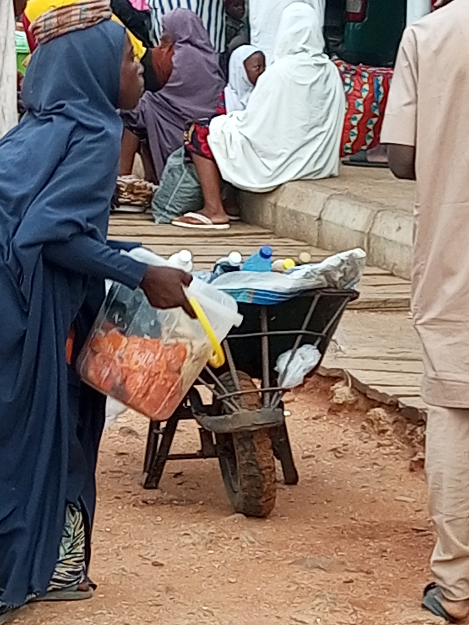 In the city of Gombe, Nigeria, a person in a dark blue hijab and patterned leggings stands on the left, holding a large transparent plastic container filled with sliced oranges. The container has a yellow handle. The person is facing right, looking away from the camera. In the background, a woman in a purple hijab sits on the ground, partially obscured by a pile of goods. Another person in a white hijab is also seated. A wheelbarrow laden with various items, including plastic bottles and a white fabric bundle, is positioned in front of these figures. The ground is sandy and uneven, with wooden planks visible in the mid-ground. The overall scene suggests a marketplace or street vendor setting.