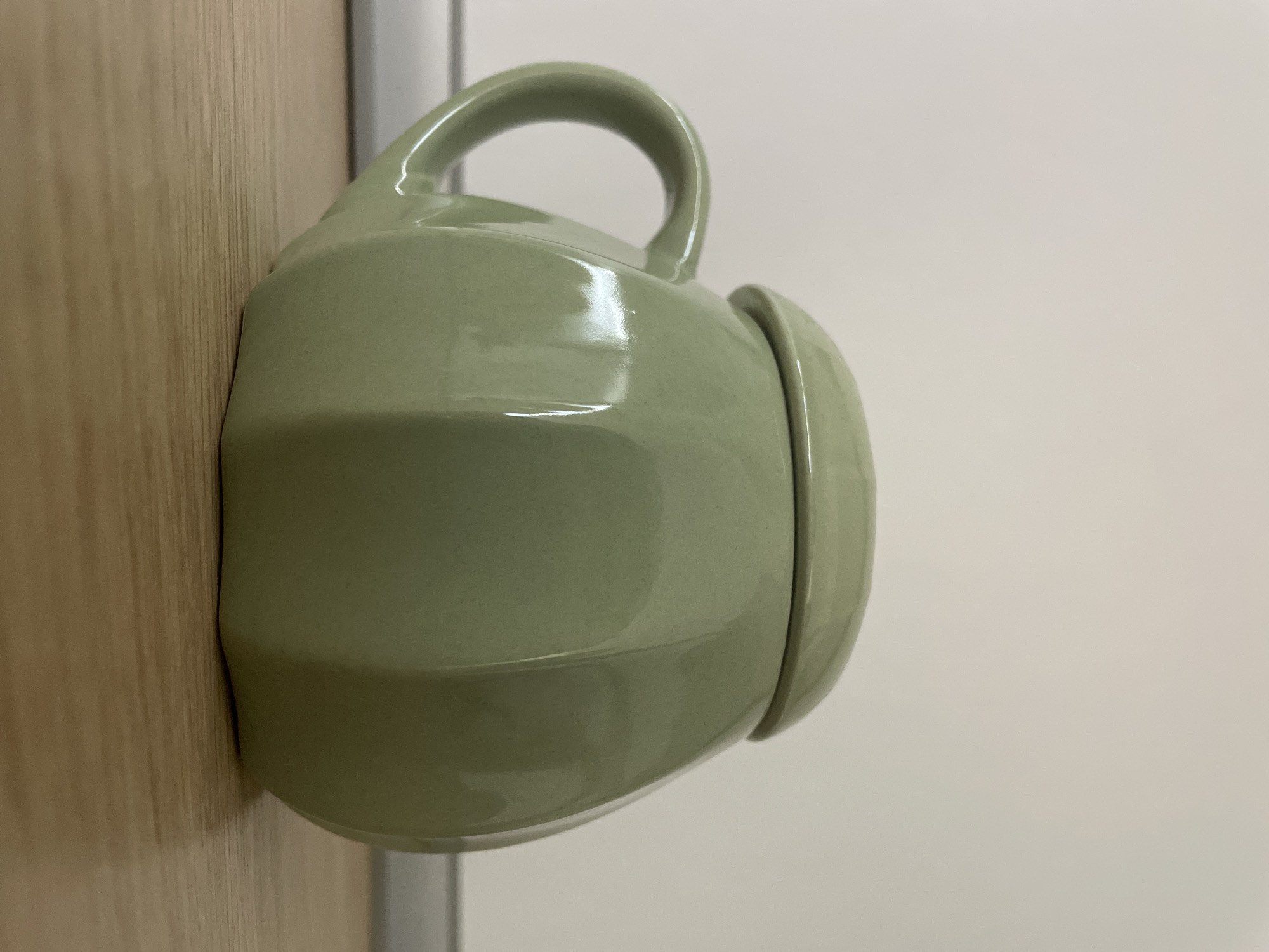A light green, faceted mug with a lid sits on a wooden surface. The mug has a handle on the left side and is oriented with its front facing the camera. The background is a plain, off-white wall. The lighting suggests an indoor environment. There are no people or animals present in the image.