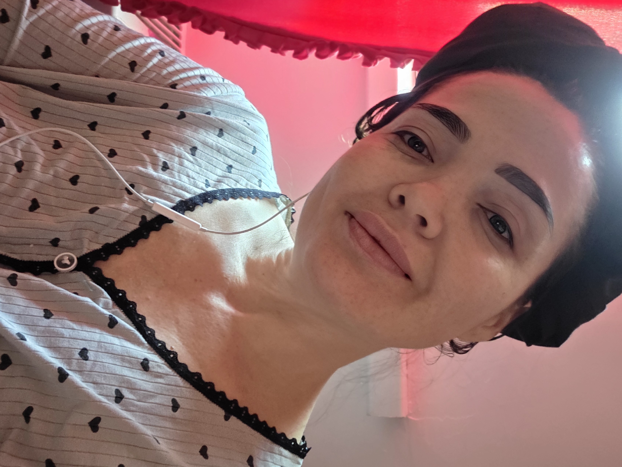A medium close-up shot shows a woman from the chest up. She is wearing a light-colored pajama top with thin vertical stripes and small black hearts. The neckline of the top is trimmed with black lace. A thin white wire from earbuds is visible near her neck. The woman has dark hair pulled back and is wearing a black head covering. Her eyes are looking towards the camera, and she has a slight smile. The background is predominantly a soft pink, with a bright red-colored area on the left side of the image, suggesting colored lighting or a draped fabric. The scene is indoor, and no specific location in Saint Petersburg, Russia, can be determined from the image.