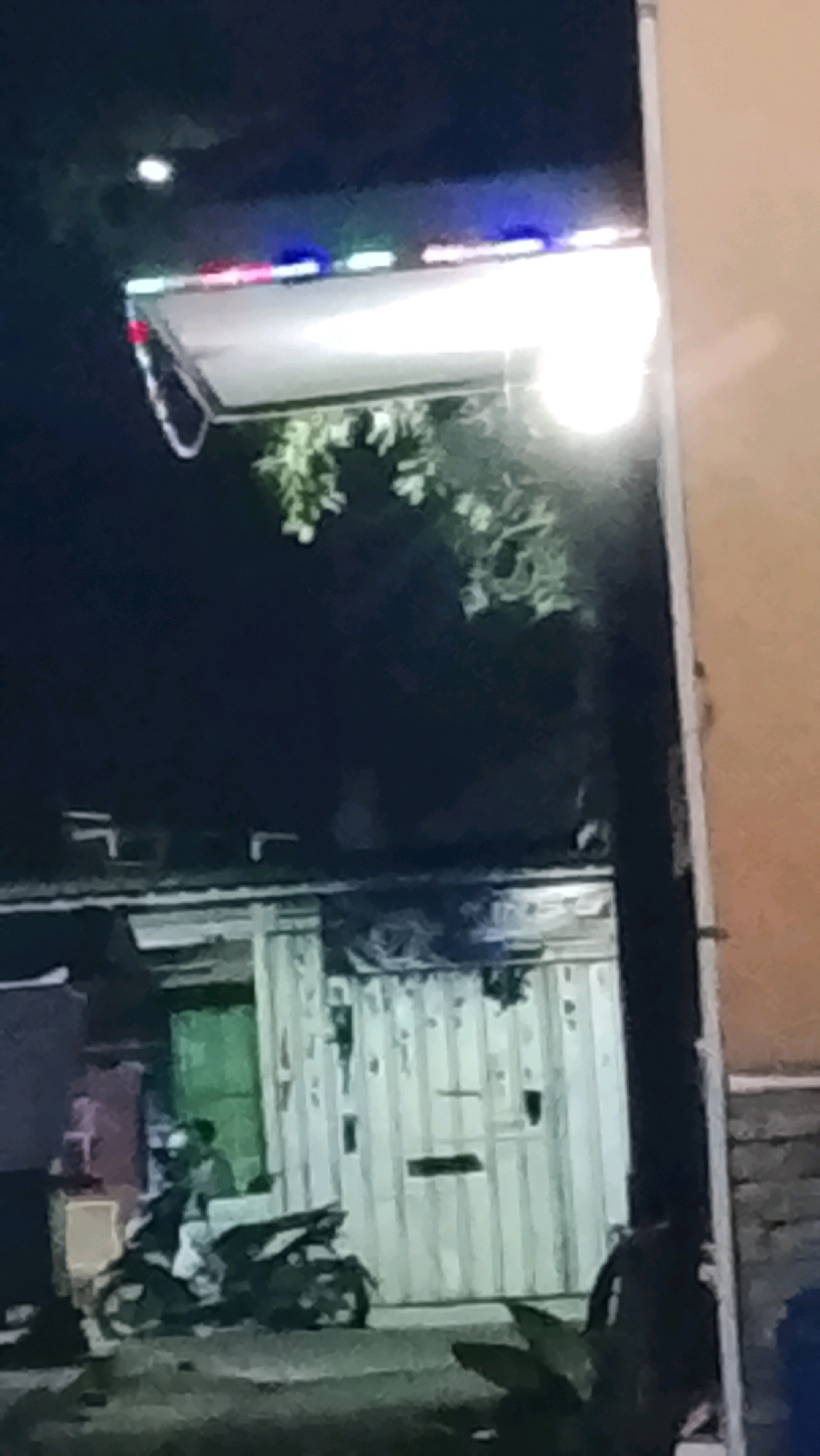 The image shows a street scene at night in Ujungrusi, Indonesia. A person is seated on a motorcycle, which is parked on the street. Behind the motorcycle, there is a building with a green door and a large corrugated metal gate. Above the building, there is a brightly lit object with red, blue, and white lights. The lighting suggests it might be a street light or a decorative light fixture. The overall scene is dark, with artificial light sources providing illumination. The area appears to be an urban or semi-urban environment.