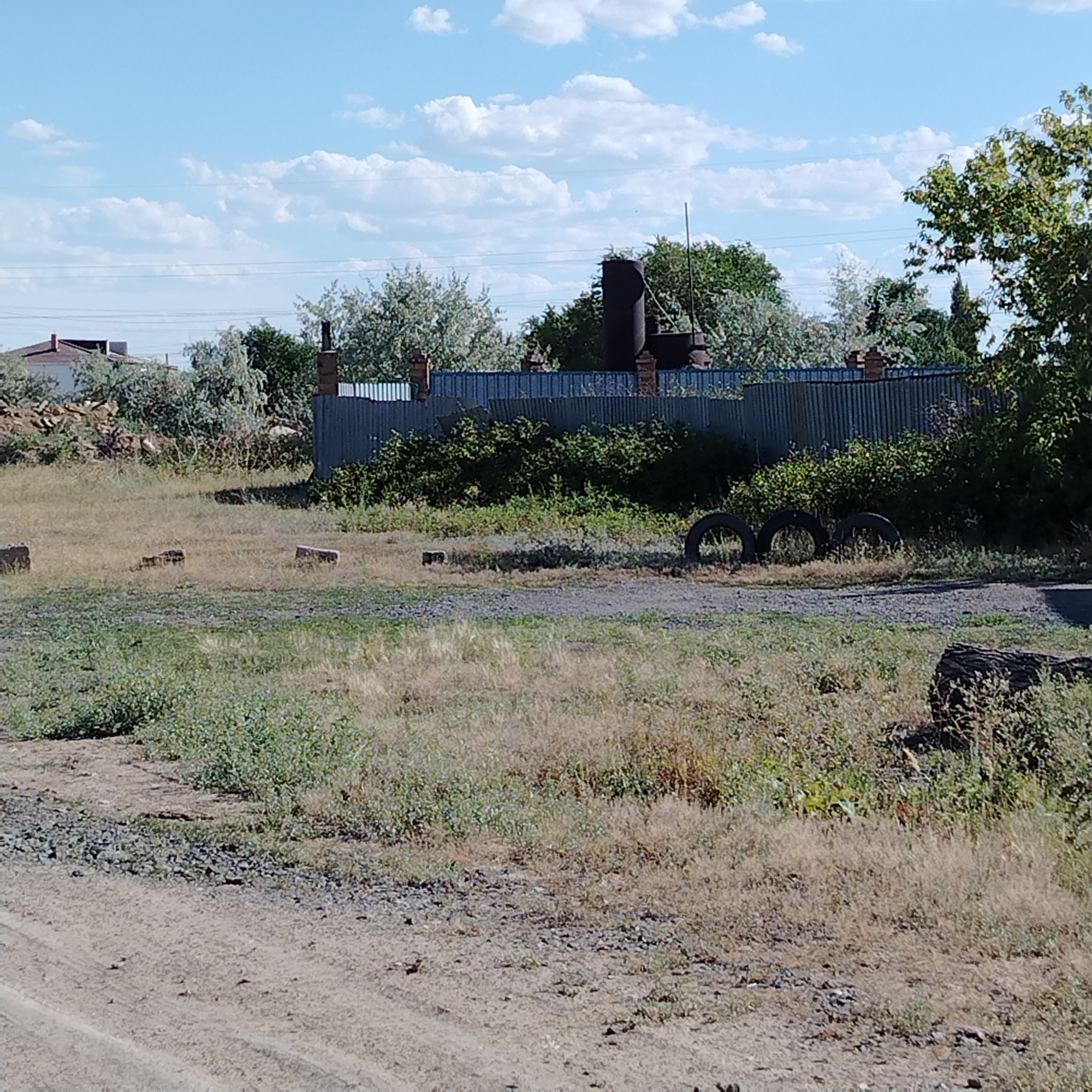 The image shows a rural or semi-industrial area with a corrugated metal fence in the background. Behind the fence, there are several tall, dark cylindrical structures, possibly industrial chimneys or silos, partially obscured by trees. There is a lot of dry grass and some green vegetation in the foreground and middle ground. Several old tires are visible on the ground. In the far background, a light-colored building with a red-tiled roof can be seen. The sky is blue with scattered white clouds. The scene appears to be located in the city of Ekibastuz, Kazakhstan, a region known for its coal mining and power generation industries, which often feature such industrial structures.