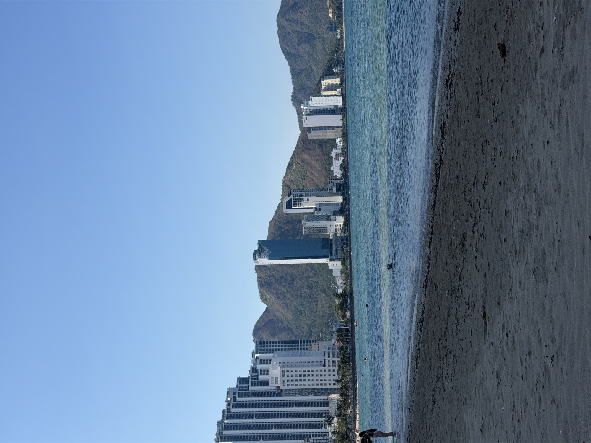 The image depicts a beach scene in Bac Nha Trang Ward, Vietnam. The foreground consists of sandy beach transitioning into a clear, blue-green ocean. In the midground, several tall buildings line the coast, backed by green, mountainous terrain. The sky is a clear, light blue. No people are clearly visible. The image captures a calm, coastal urban environment.