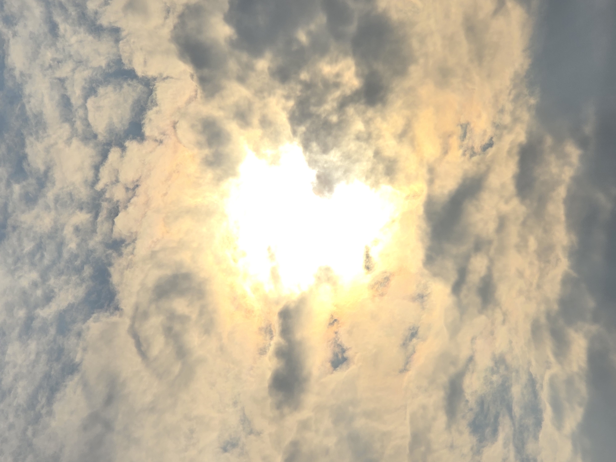 The image shows a cloudy sky with the sun shining brightly through the clouds. The clouds are white and gray, with some areas of blue sky visible. The sun is a bright, white orb in the center of the image, with rays of light emanating from it. The overall impression is one of a slightly overcast but bright day. The image is a photograph taken from below, looking up at the sky. There are no people, animals, or objects visible in the image. The location is described as Ермолаево, Russia, which is a city. However, the image itself does not contain any specific visual cues to confirm this location.