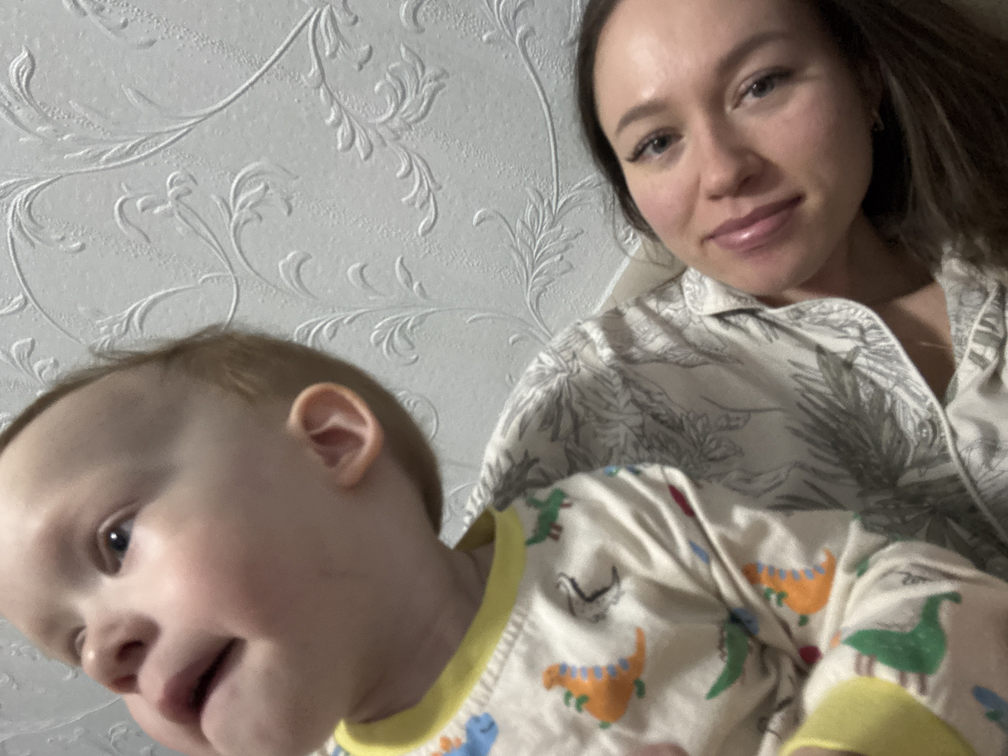 This close-up indoor image features an adult woman and a baby, likely a mother and child. The woman, positioned in the upper right, has long dark hair and a gentle smile, looking directly at the camera. She is wearing a light-colored top with a subtle, leafy pattern. The baby occupies the lower left foreground, seen mostly in profile with a soft, content expression and light hair. The baby is dressed in a light-colored sleepsuit or pajamas adorned with colorful cartoon dinosaurs (orange, blue, green). The background is a light-toned wall covered in wallpaper featuring an embossed or raised floral/vine pattern. The photo captures an intimate and joyful moment between the two subjects, possibly a selfie. The pajamas suggest a relaxed home setting, likely in Ферма, Russia. No specific time of day or weather is discernable, and there is no visible text.