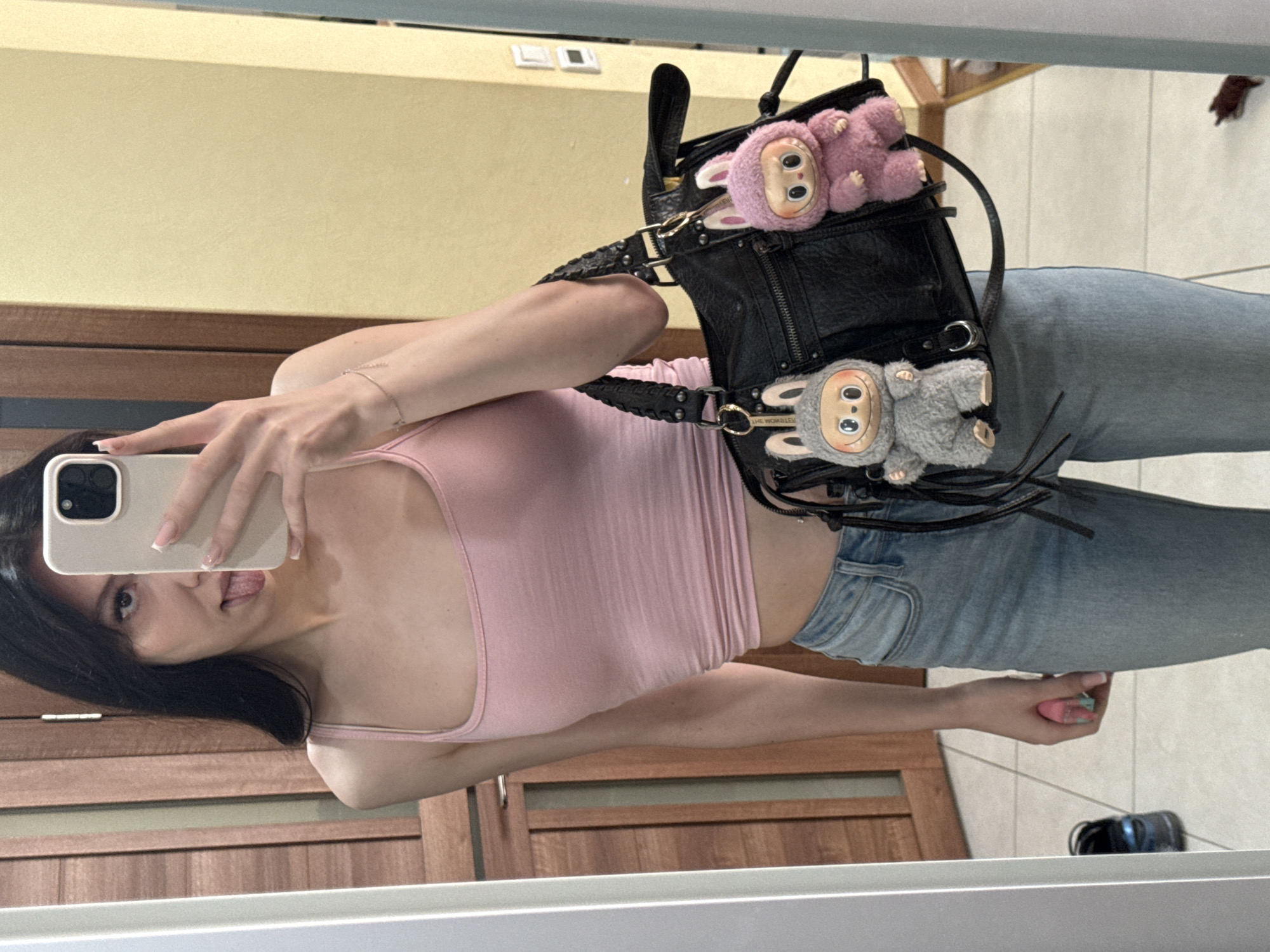 A young woman with dark hair is taking a mirror selfie in what appears to be a bedroom or dressing area. She is wearing a pink crop top and light blue jeans, and she is holding a black handbag with two furry stuffed animal keychains. The woman is sticking her tongue out at the camera. The background shows a wooden door and a yellow wall. The floor is tiled, and a pair of black shoes are visible near the bottom left of the frame. A brown object, possibly a toy or a pet, is on the floor in the lower right corner. The overall scene suggests a casual, personal setting. There is no specific information in the image to suggest a location in the city of Khimki, Russia.