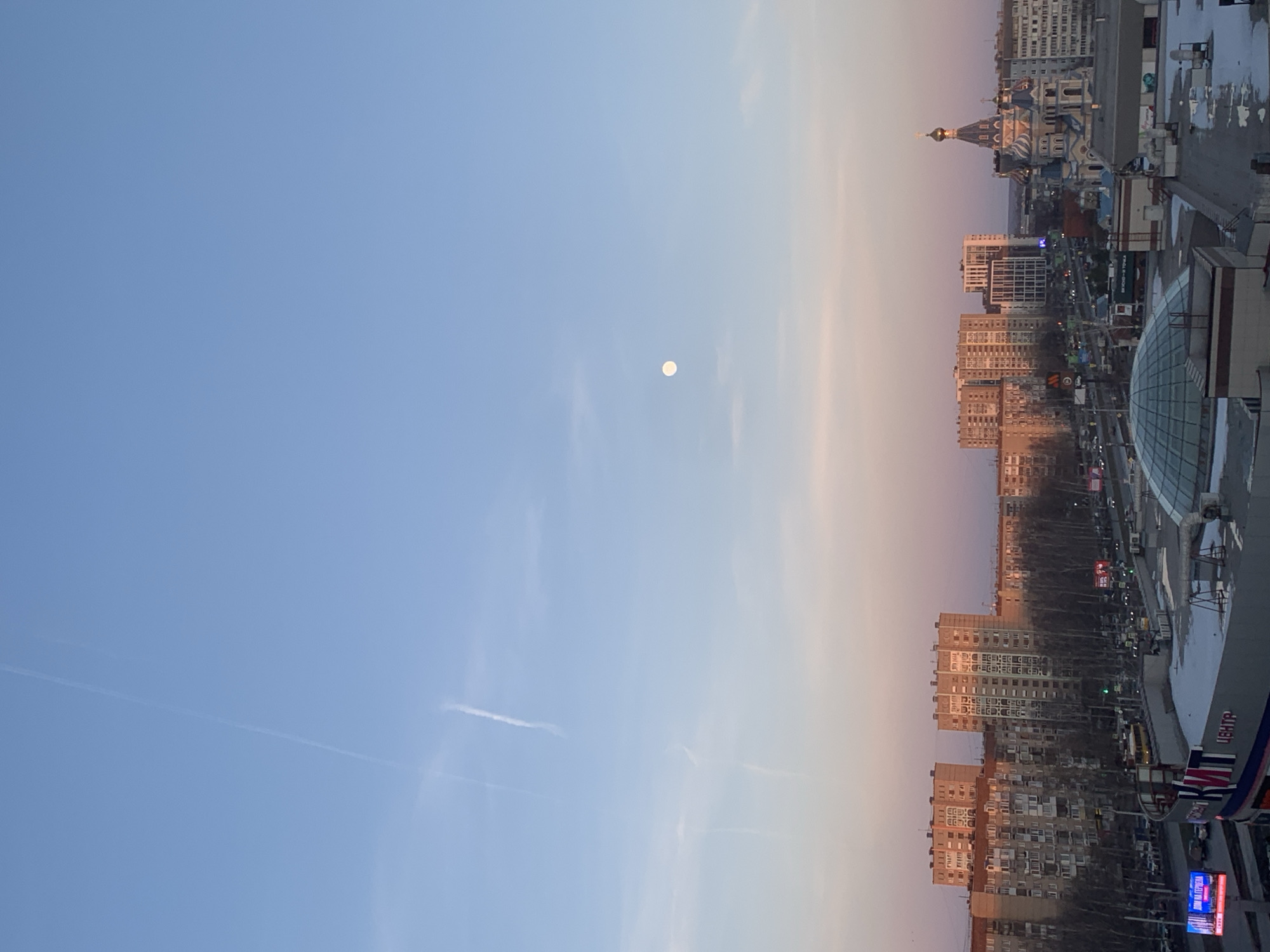 The image captures a cityscape in Izhevsk, Russia, under a pale blue and peach-colored sky. The full moon is visible high in the sky. The buildings range in height and architectural style, including a domed cathedral with golden spires, high-rise apartment complexes, and a modern shopping center with a glass dome. Street lights and traffic signals are visible amongst the buildings. Some rooftops show patches of melting snow. The image is taken from an elevated perspective, possibly from a high-rise building.