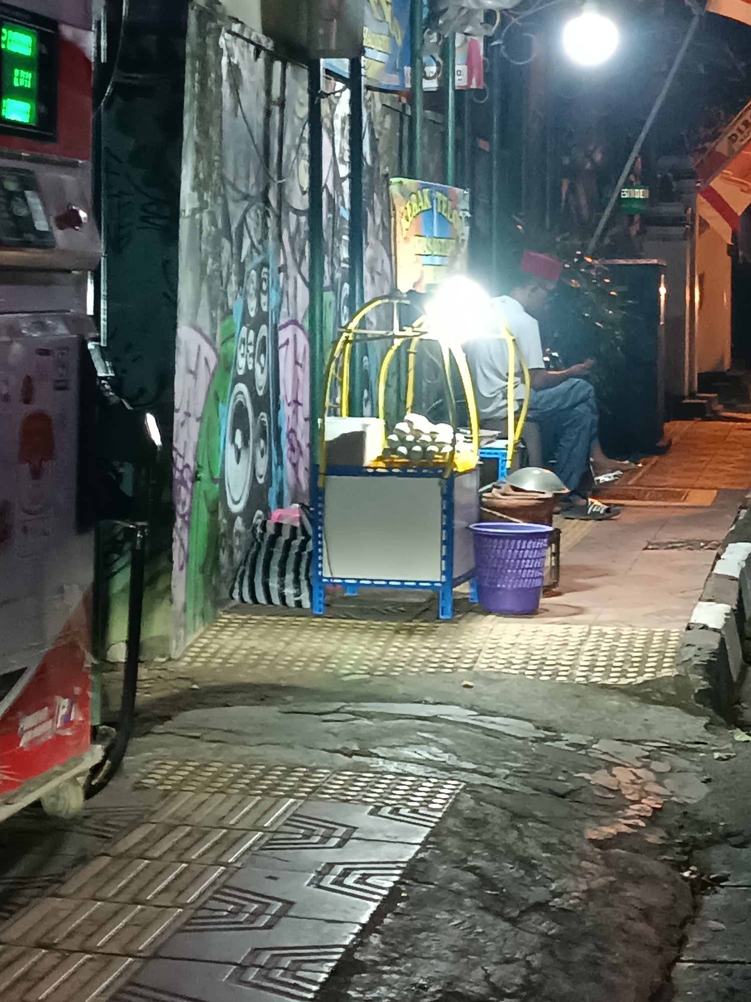This is a nighttime photograph of a street vendor in Yogyakarta, Indonesia. The scene is illuminated by a bright overhead light. In the foreground, a patterned pavement leads to a dimly lit street. To the left, a red vehicle with a green digital display is partially visible. In the center, a food cart is set up with a yellow metal frame and a white board. Several items, possibly food, are displayed on the cart. A person wearing a white t-shirt and blue pants is seated behind the cart, appearing to be using a mobile phone. A purple trash bin and a brown container are also present. The background features a wall covered in graffiti. The overall atmosphere is that of a busy urban street at night.