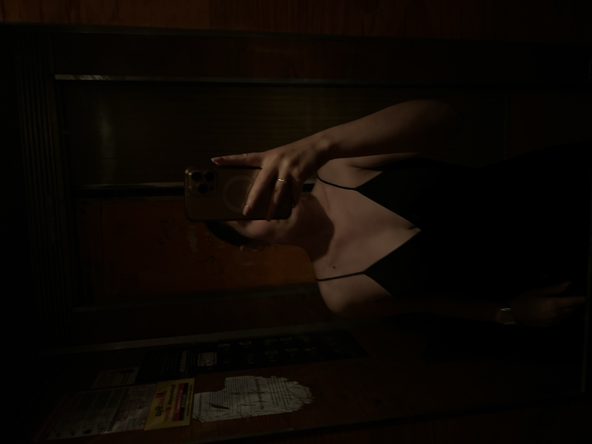 The image is a low-light, vertically oriented, reflected selfie taken in what appears to be an elevator. The main subject is a person, visible from the shoulders up, holding a smartphone in their right hand to take the picture. The person is wearing a black dress with thin spaghetti straps. A thin, gold-toned ring is visible on the ring finger of the hand holding the phone.
The background is dark, with wooden paneling visible at the top and sides, suggesting the interior of an elevator cabin. Reflections in the elevator's mirrored surface reveal details of the surroundings, including what looks like signage or posters on the elevator wall or doors. One sign, partially obscured and with text in Ukrainian, is visible in the lower left section of the image.
The lighting is dim, creating a dramatic and somewhat mysterious atmosphere. The time of day is indeterminate due to the indoor setting and low light. No specific weather conditions are suggested. The setting is an elevator in Ternopil, Ukraine, as indicated by the provided location context. The image conveys a sense of personal reflection or a private moment captured through a selfie.