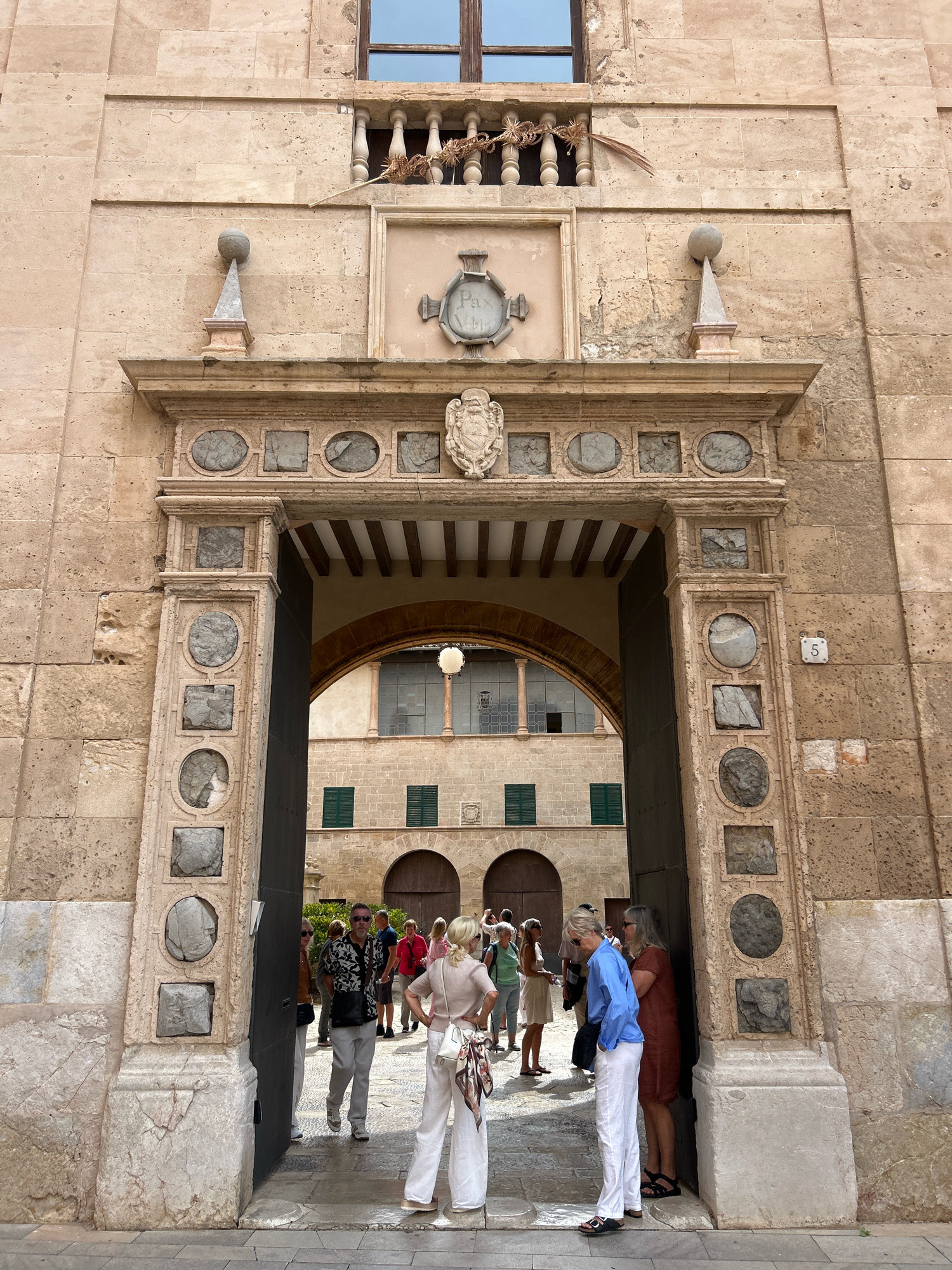 The image shows the entrance to the Palau de l'Almudaina in Palma de Mallorca, Spain. The ancient stone building features an ornate archway adorned with decorative carvings and circular stone inlays. A group of tourists, dressed in casual summer attire, are gathered at the entrance, some peering into the courtyard beyond. The courtyard itself is visible through the archway, revealing more stone architecture and greenery. The scene is bathed in daylight, casting shadows that accentuate the building's textures and details. The overall impression is one of historical grandeur and the presence of modern visitors exploring the site.
