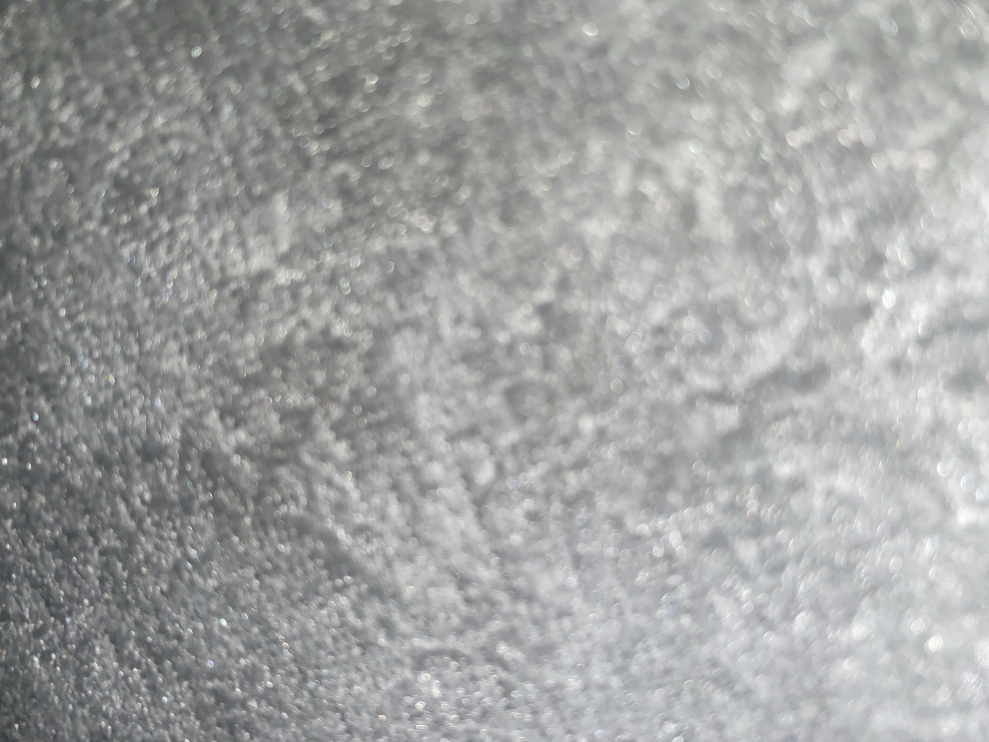 The image is an abstract, out-of-focus shot of a shimmering, granular surface, likely glitter or tiny reflective particles. The colors are predominantly shades of gray and white, with subtle hints of iridescence. There are no discernible objects, people, or environmental features that would allow for identification of a specific location, including Ablanitsa, Bulgaria. The texture appears to be densely packed and uniform across the frame.