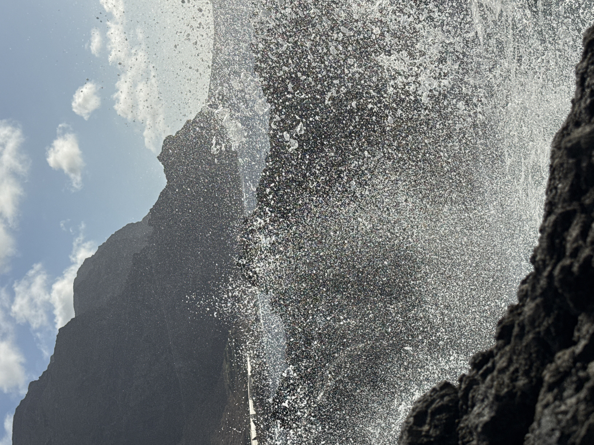 The image captures a dramatic scene of waves crashing against rocky shores under a partly cloudy sky. In the foreground, dark, rugged rocks are visible, with a large portion of the frame filled with a dense spray of white water droplets, suggesting a powerful wave impact. In the midground, the dark, rocky coastline continues, with the sea surface appearing turbulent. In the background, a dark, imposing cliff face rises against a bright blue sky dotted with white clouds. The overall impression is one of raw, natural power and a dynamic coastal environment. The specific location is Unknown, Unknown.
