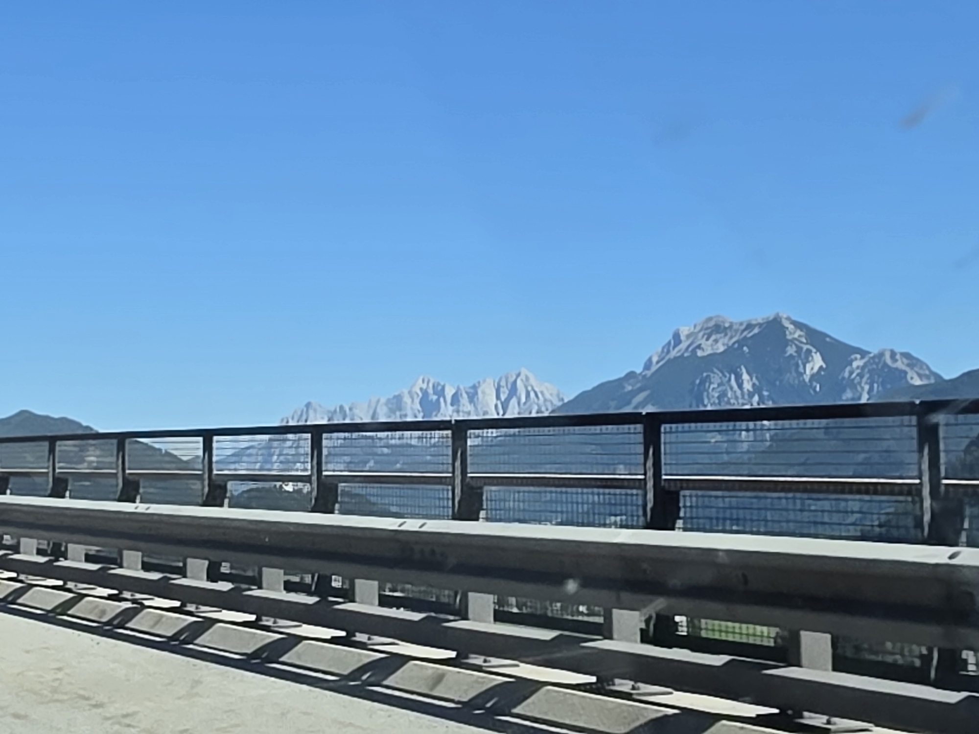 The image shows a view from a bridge with a metal and glass barrier. In the background, there are large, rugged mountains under a clear blue sky. The mountains appear to be snow-capped in some areas. Below the barrier, there is a glimpse of greenery and a road surface. The scene suggests a highway or elevated road passing through a mountainous landscape. The specific location in Ardning, Austria, is not directly identifiable from the visual elements in the image alone, but the mountainous terrain is characteristic of the Austrian Alps.