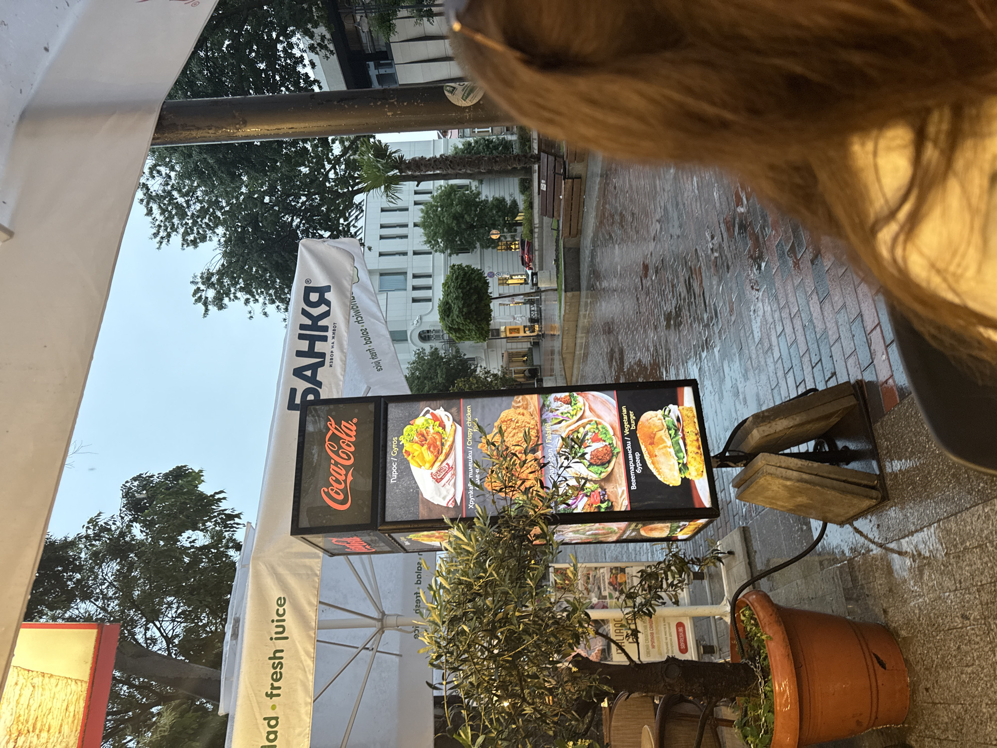 The image captures an evening street scene in Varna, Bulgaria, viewed from a first-person perspective, likely from under an awning or umbrella. A person's brown hair is visible in the top right quadrant, partially framing the view.
In the midground, a prominent, vertically oriented, illuminated menu sign advertises various food items. The sign features a "Coca-Cola" logo at the top. Below it, pictures and text display "Tupoc / Gyros," "Хрупкав пилешки / Crispy chicken," "Falafel," and "Вегетариански / Vegetarian burger." Below the menu, an olive tree in a large terracotta pot stands on the wet pavement. White awnings or large umbrellas are also visible, one partially obscured in the foreground, and another with text reading "ad • fresh juice." To the left, a large white banner with Cyrillic text "БАНКЯ" (BANKYA) and "ИЗВОР НА ЖИВОТ" (SOURCE OF LIFE) is visible.
The background shows a wet street or sidewalk paved with bricks, reflecting the ambient light. This indicates recent rainfall. Beyond the street, multi-story buildings, including a prominent white one, are visible. Trees, including a palm tree, line the street. A dark street pole and a wooden bench are also present. The overall atmosphere is that of a bustling urban evening after a rain shower. Other partially visible text includes "SOFRESH.BG," "ITALIANC," "СВЕЖА САЛАТЕН," and "КРУПКАВ САНДА."