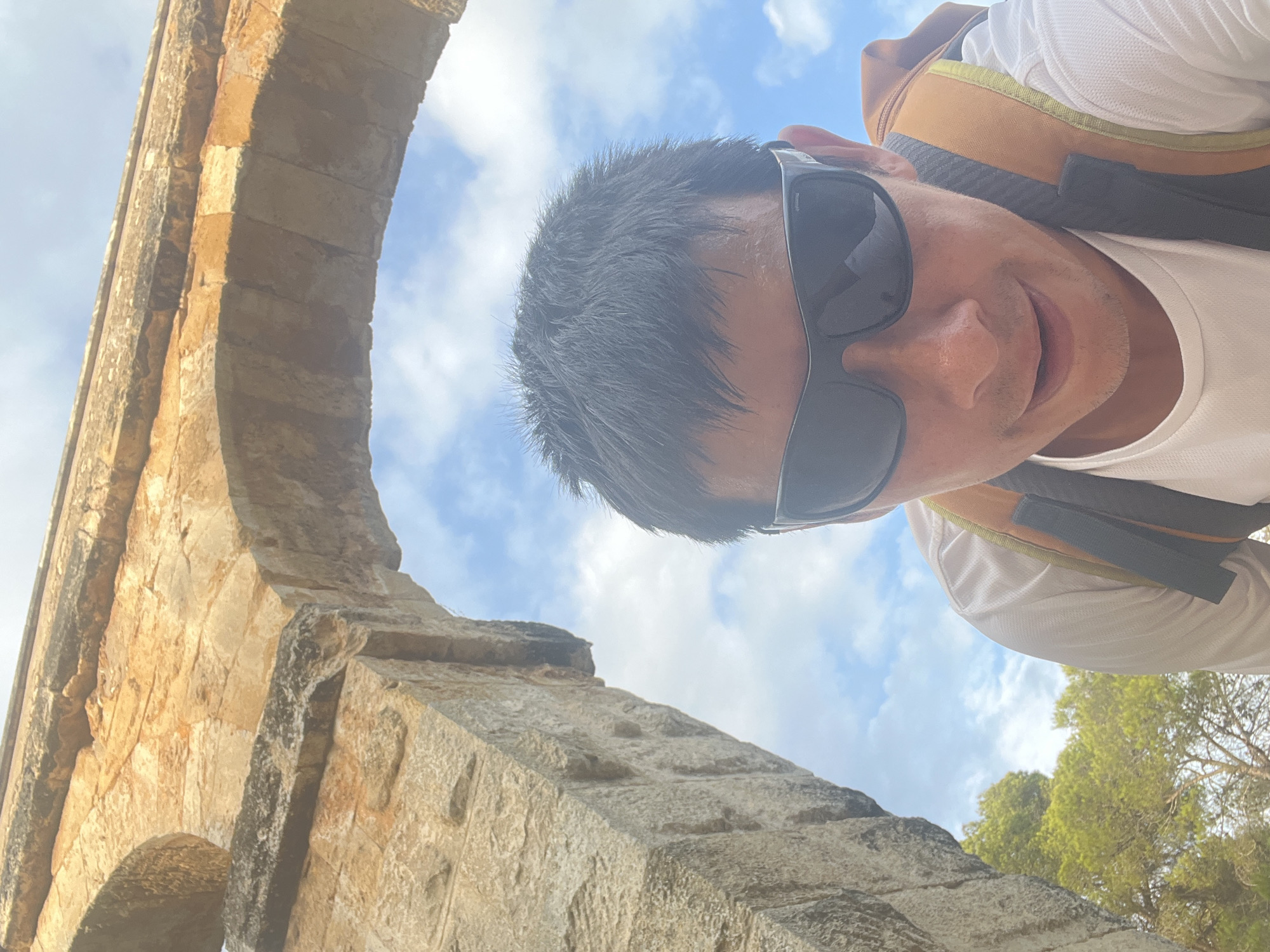The image is a selfie taken from a low angle, looking up towards a stone aqueduct. A person, wearing sunglasses and a white t-shirt with a backpack, is in the foreground. The aqueduct appears to be an ancient Roman structure, possibly part of the Pont del Diable in Tarragona, Spain. The sky is blue with scattered clouds, and trees are visible in the background. The lighting suggests it is daytime, possibly late morning or early afternoon.