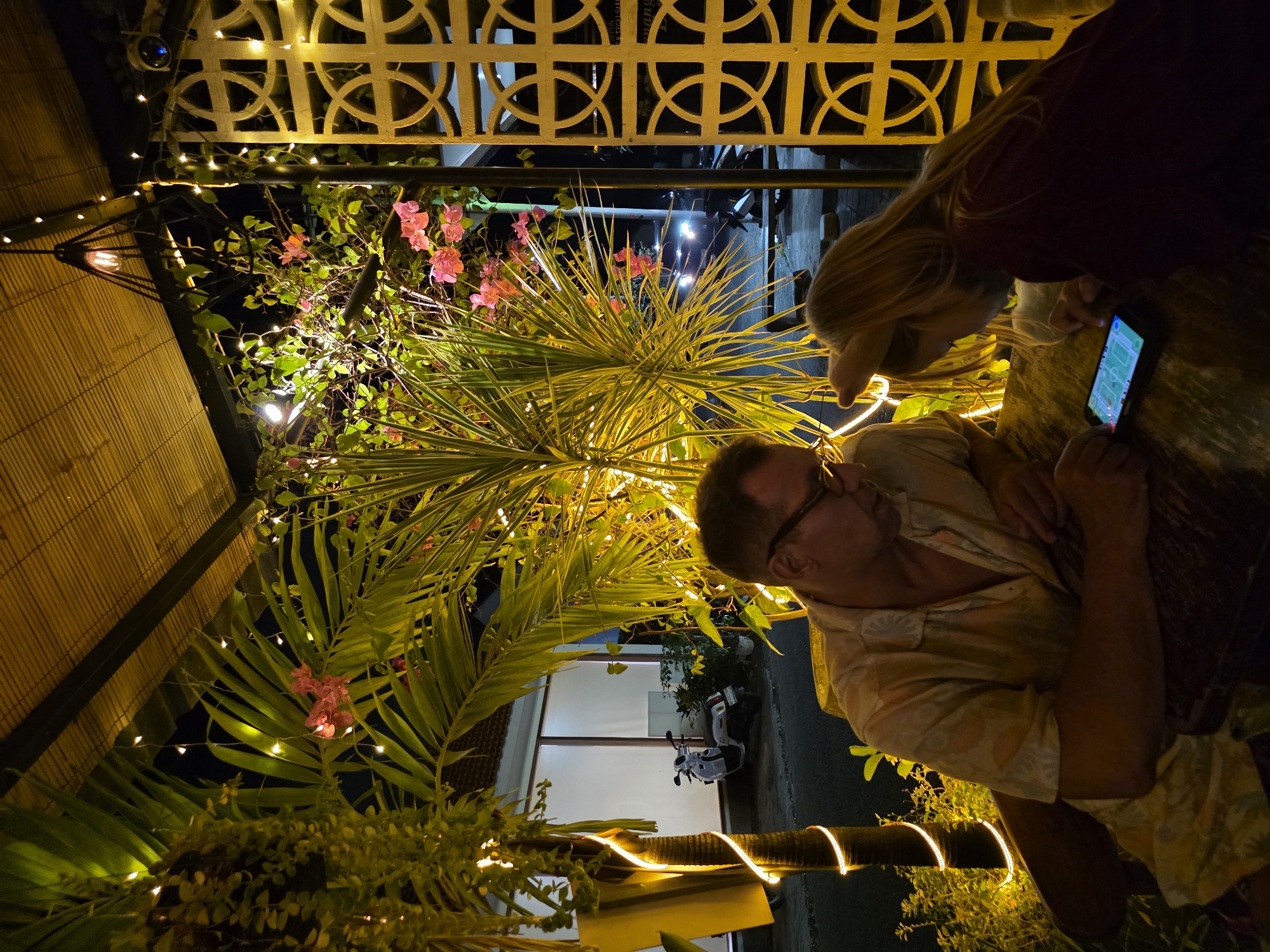 The image captures an evening scene at an outdoor dining establishment in North Kuta, Indonesia, likely a restaurant or cafe. Two individuals, an adult male and a child, are seated at a table, engrossed in a smartphone. The male, wearing glasses and a light-colored patterned shirt, leans forward, his arm resting on the table. The child, with blonde hair and wearing a dark top, is focused on the phone's screen, holding a finger over it.
The environment is characterized by lush tropical foliage, including palm fronds and flowering plants, illuminated by warm, golden string lights. A bamboo-thatched roof covers part of the area, with a distinctive decorative metal screen on the right side. In the background, a white scooter is parked near a building with white walls and a tiled roof. The lighting creates a cozy and somewhat intimate atmosphere, despite the presence of the outdoor elements and passing vehicles visible in the distance.