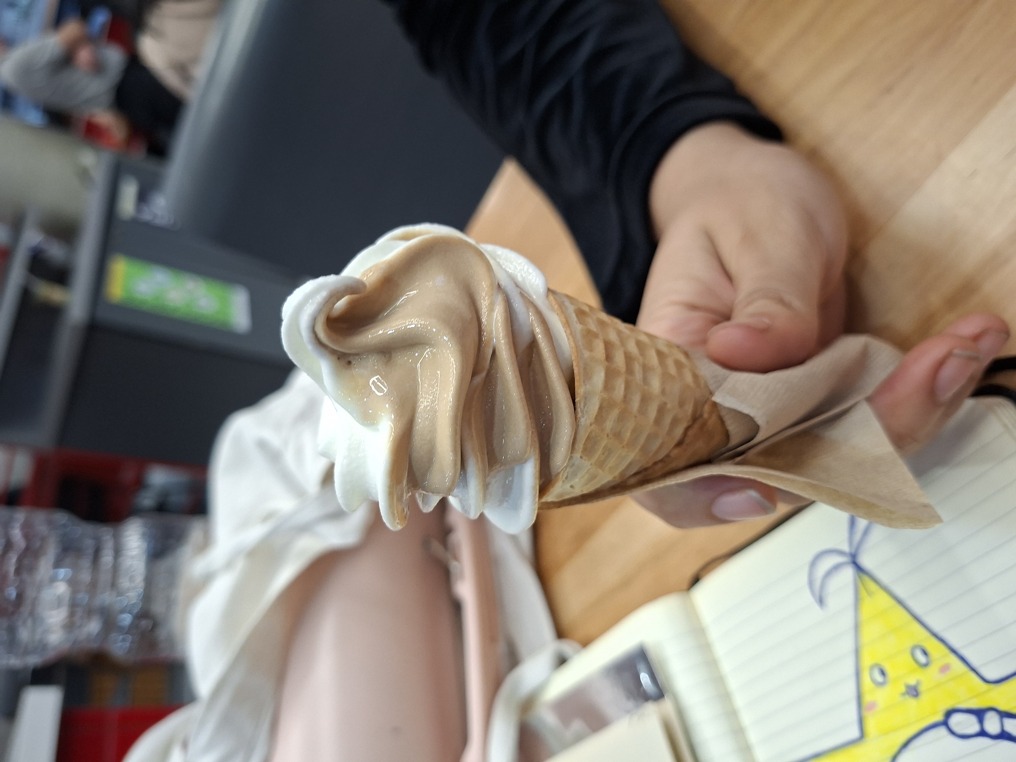 A close-up shot shows a hand holding an ice cream cone, predominantly vanilla and dulce de leche swirl, wrapped in a tan paper napkin. The cone rests partially on an open notebook, which has a drawing of a yellow star with a party hat. A wooden-patterned table surface serves as the immediate background. The person holding the cone is wearing a black long-sleeved shirt. Soft focus elements in the background suggest an interior space; a beverage cooler or refrigerator with a partially visible brand name. No explicit indicators tie this to a specific location, such as Buenos Aires, Argentina.