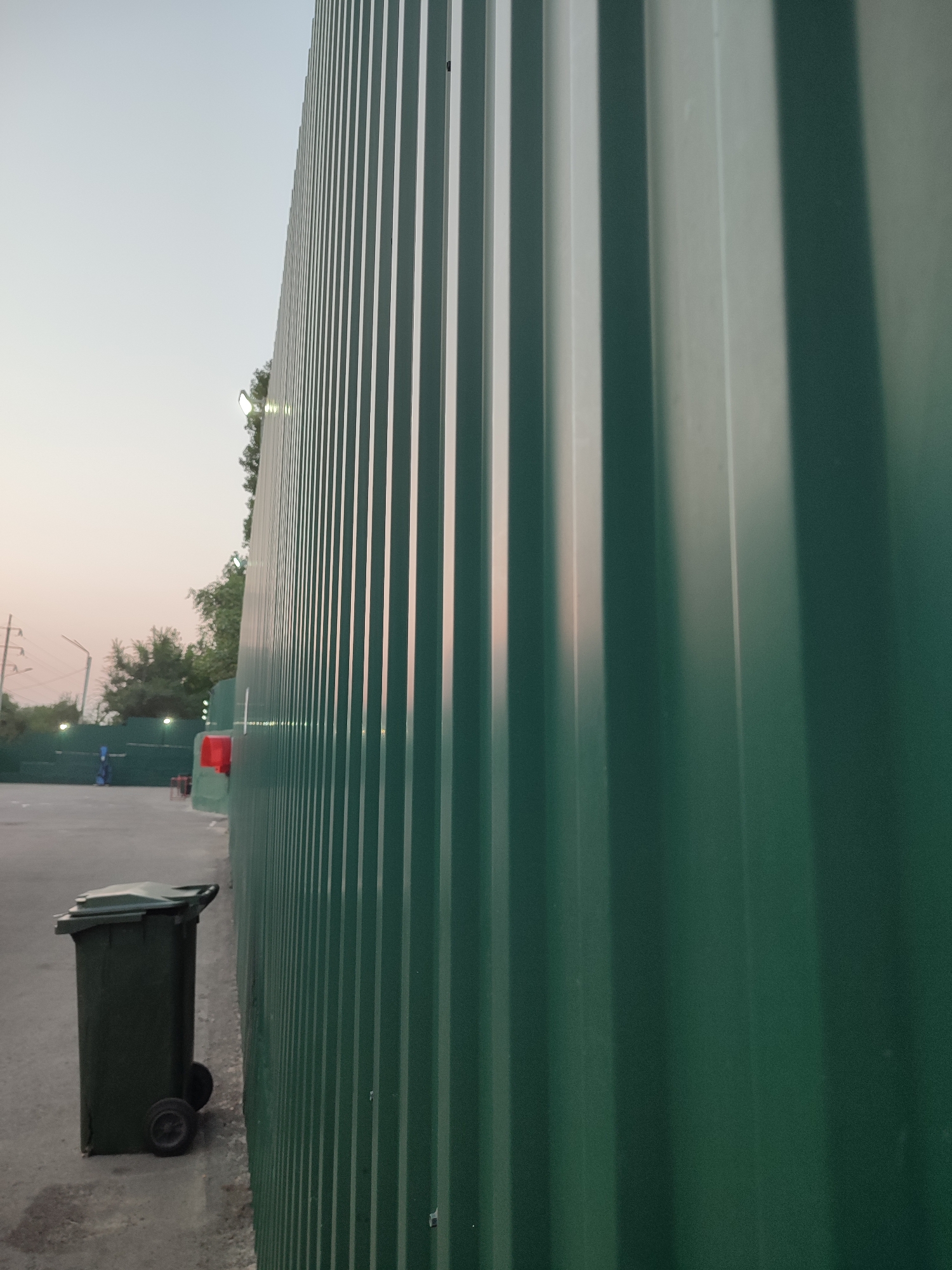 The image captures an outdoor scene, with a prominent green corrugated metal fence dominating the right side of the frame. To the left of the fence, a grey concrete surface is visible, with a dark green wheeled trash bin positioned near the bottom left corner. In the background, beyond the fence, trees and some structures, including what appears to be a red object, are partially obscured. The sky above is a pale, hazy blue, suggesting either early morning or late evening. The overall impression is of an industrial or service area. The location is not specified in the image.
