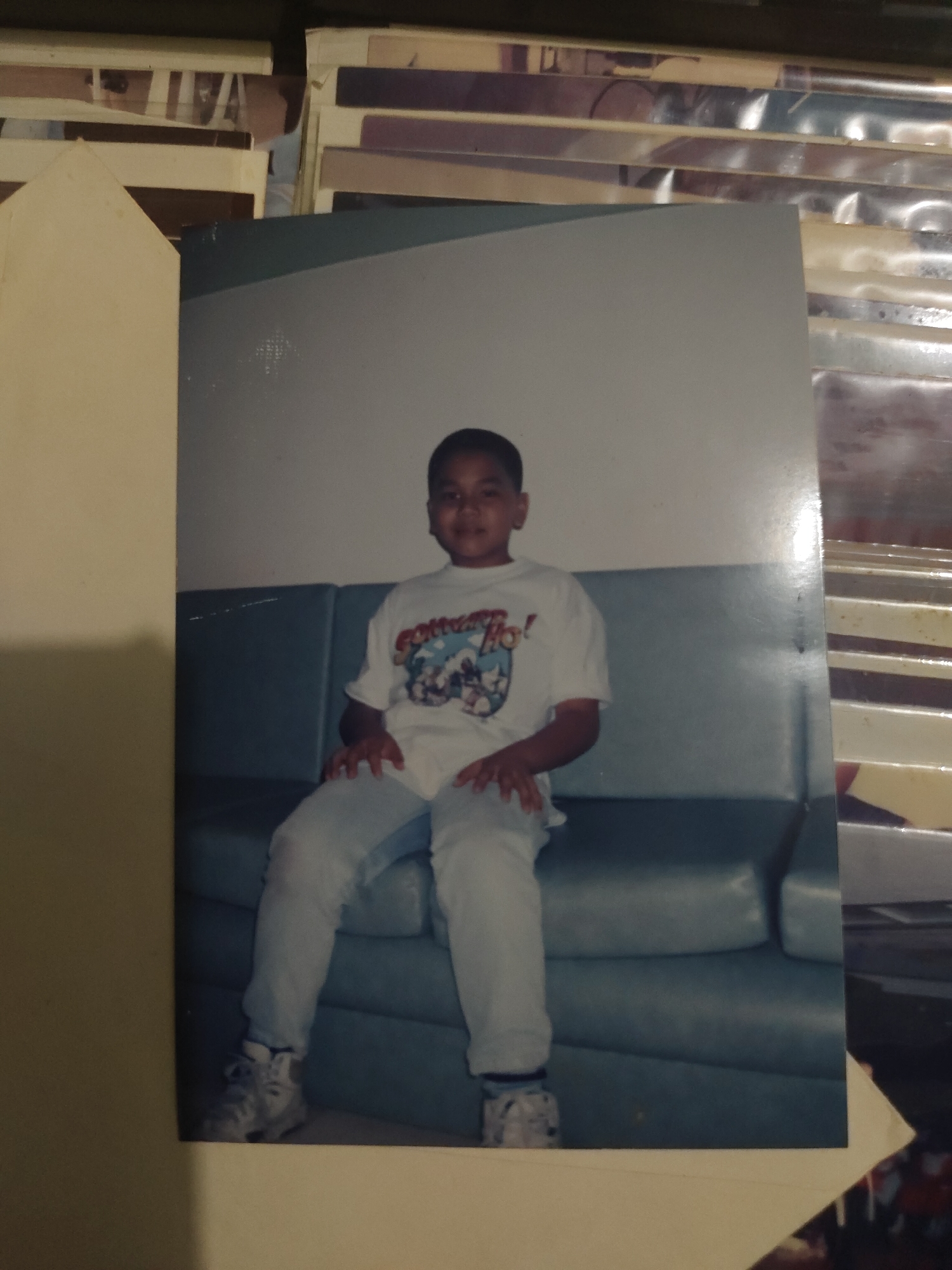 An undated photograph depicts a young male child seated indoors in an unknown city and state. The child, appearing to be approximately 5-8 years old with short, dark hair, is positioned centrally on a blue-grey upholstered sofa or bench. He wears a white short-sleeved t-shirt featuring a graphic design that includes animated characters and text, partially legible as "GOTTA GO!". His lower attire consists of light blue pants or jeans, white socks, and white sneakers with dark accents. The child's hands are resting on his lap, and he is looking directly towards the camera with a neutral to slightly smiling expression. The background consists of a plain, light-colored wall above the sofa. The physical photograph itself is framed on the left by a light beige or cream-colored element, possibly a page from a photo album, and the upper right background shows stacks of other photographs or photo albums, partially covered in clear protective sleeves, creating a layered effect around the main image.