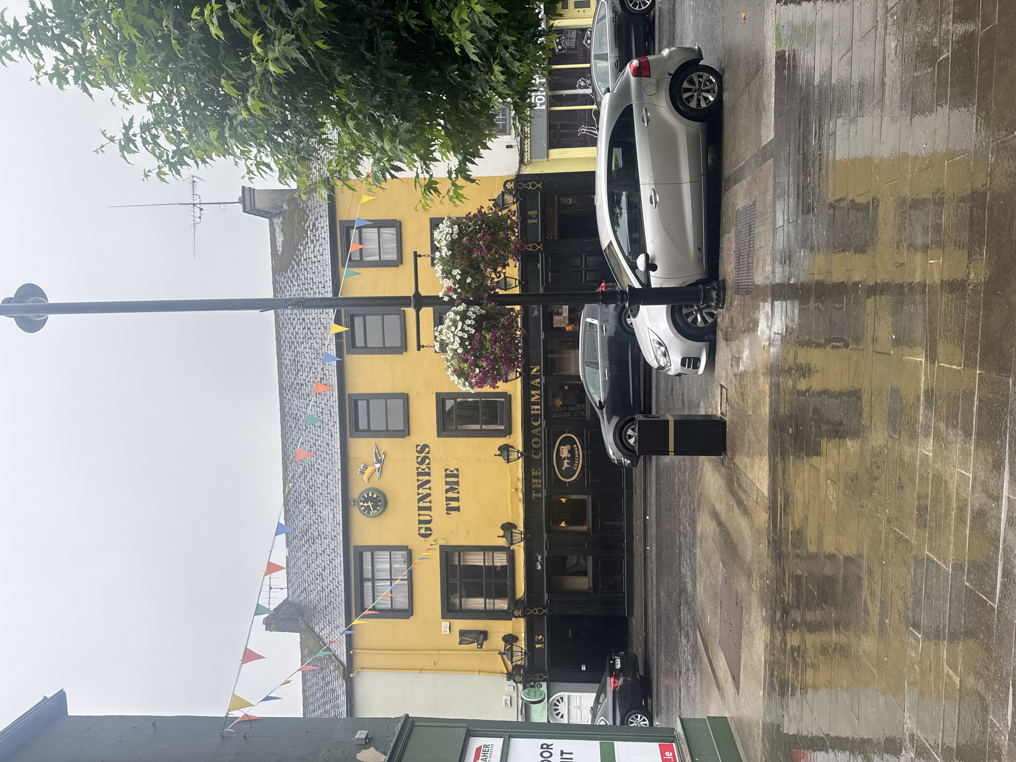 The image shows a street scene in Clonmel, Ireland, on a rainy day. A bright yellow building with "GUINNESS TIME" written on it, a clock, and a toucan logo is the prominent feature. The building appears to be a pub called "The Coachman." Bunting in various colors is strung across the street. Cars are parked on the street and the pavement is wet, reflecting the surrounding buildings and the sky. There are hanging flower baskets in front of the pub and a lamppost. A dark grey car is parked on the left, a silver car is in the foreground, and a dark grey SUV is parked on the right. A black trash bin is also visible. The overall atmosphere is damp and overcast, characteristic of Irish weather.