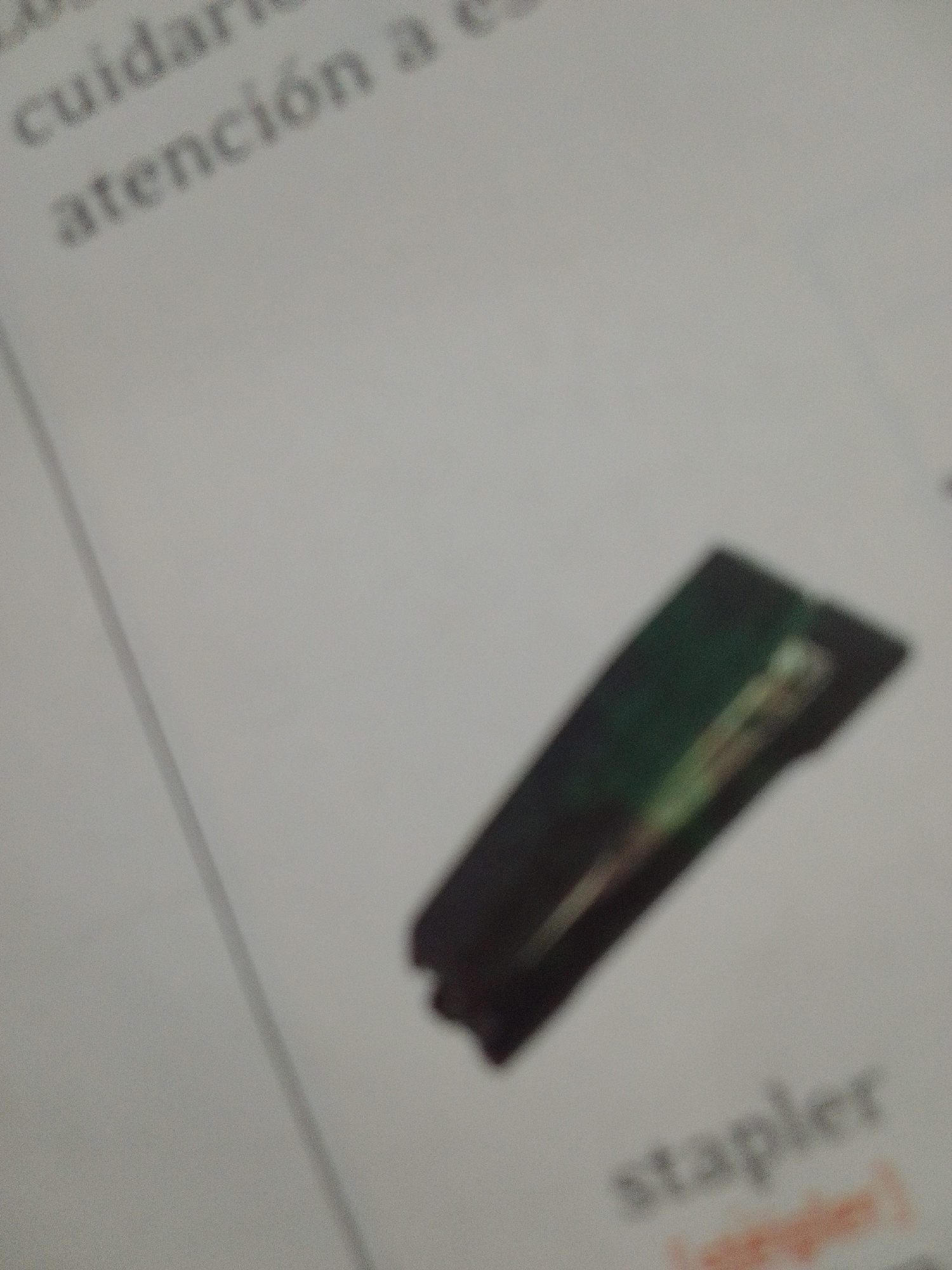 The image shows a close-up, slightly out-of-focus view of a page with text and an object. At the top of the page, text in Spanish is partially visible, including the words "cuidar" and "atención a". In the lower right quadrant, a dark-colored object, likely a stapler, is present, with some reflective highlights suggesting it is made of plastic or metal. Below the stapler, the word "stapler" is printed in English. The background is a plain, light-colored surface, possibly paper or a table. There is no indication of the image being located in the city of La Tagua, Venezuela.