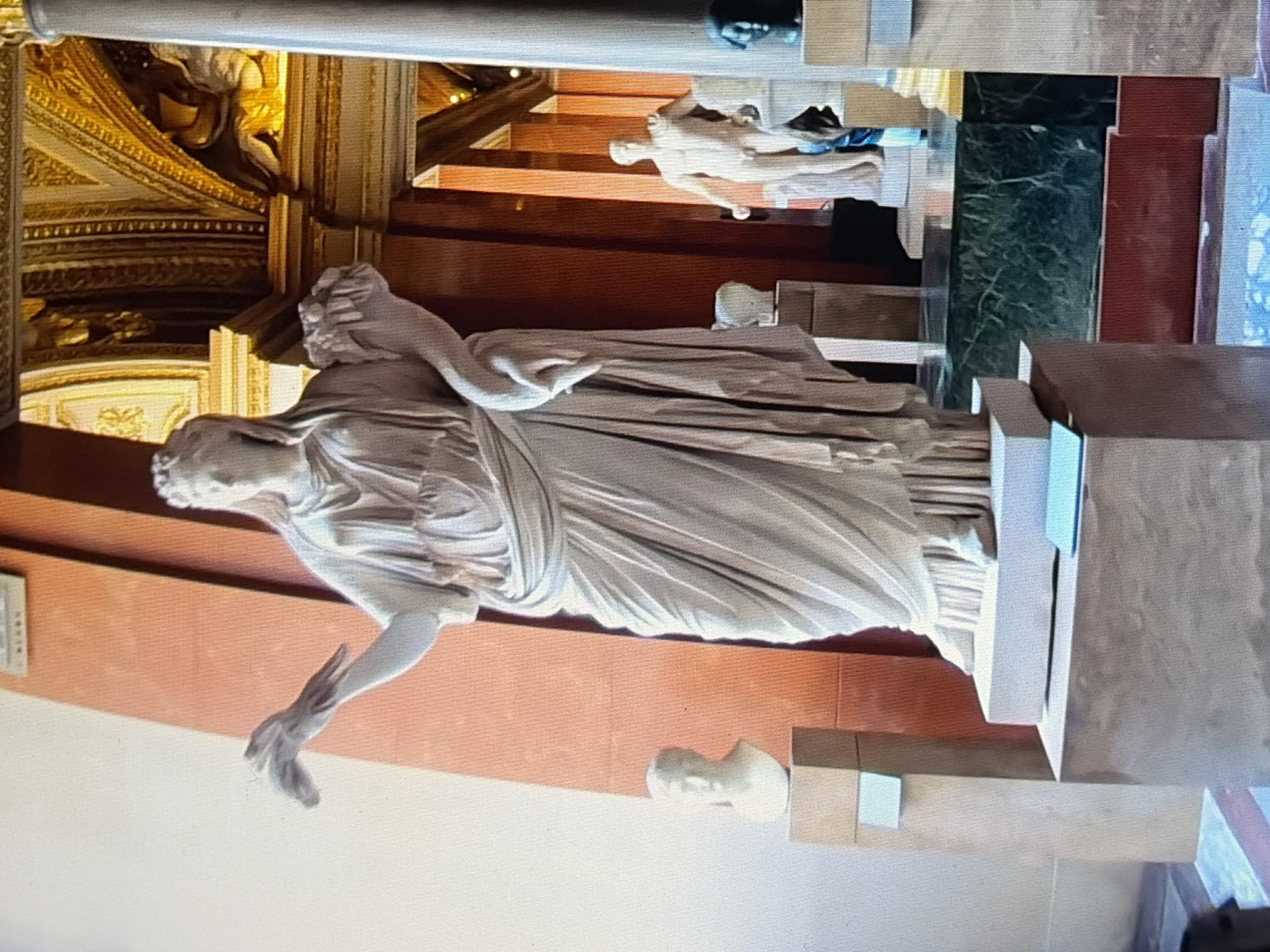 A full-body marble statue of a woman stands prominently in the center of the frame, adorned in a draped garment. Her left arm is bent, holding a bouquet of flowers, while her right arm is extended, holding what appears to be a bird. The statue is positioned on a pedestal, which rests on a larger, multi-tiered base. To the left of the main statue, a smaller marble bust of a man is displayed on a pedestal. In the background, another marble statue of a nude male figure is visible, along with architectural elements of a grand interior space, including columns, ornate gold detailing on the ceiling, and warm-toned walls. The scene is illuminated with bright lighting, suggesting an indoor exhibition space like a museum.