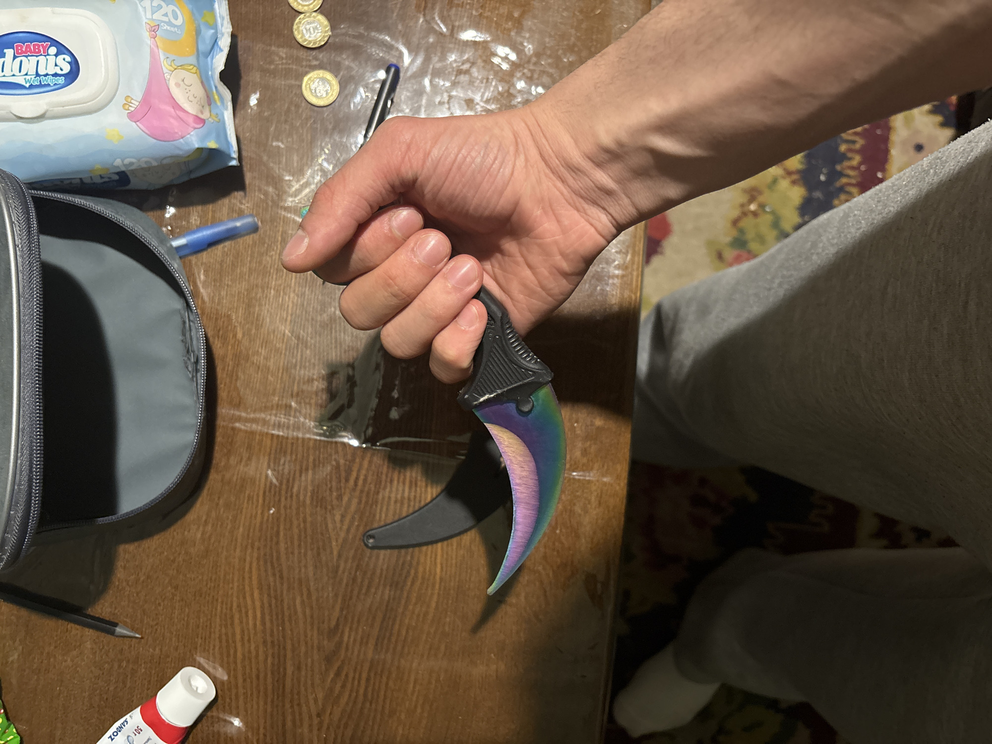 A man is holding a karambit knife with a rainbow-colored blade. The background shows a wooden table with various items, including baby wipes, coins, a pen, a pencil, a tube of ointment, and a gray zipper bag. The man is wearing gray sweatpants. The location is not identifiable as Yerevan, Armenia.