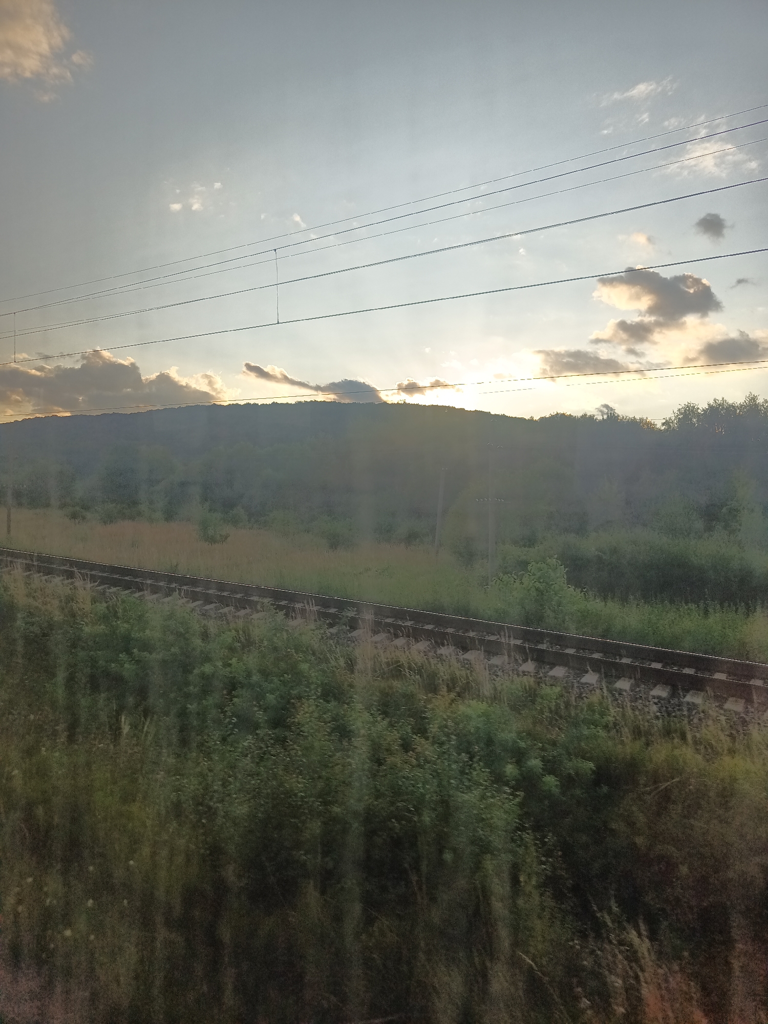 The image shows a rural landscape with a train track in the foreground. A tree-covered hill is visible in the background, with the sun setting behind it. The sky is partly cloudy, with the sun casting a warm glow. The overall scene is peaceful and serene.
