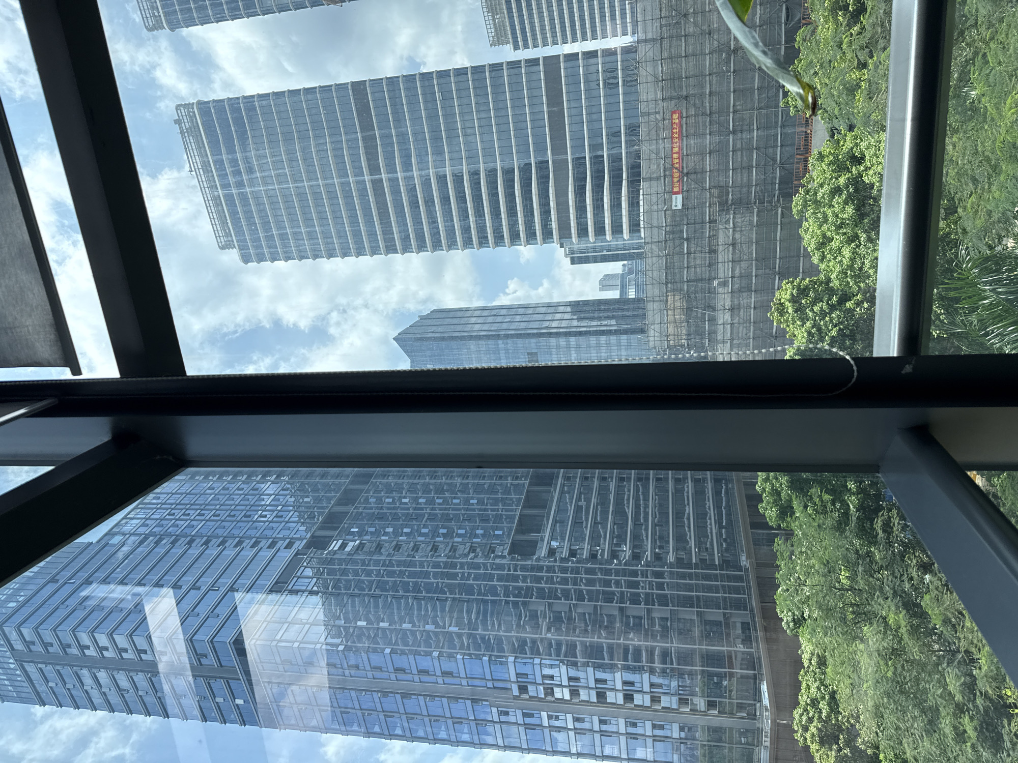 This is a photograph taken from inside a building, looking out through a window frame at a cityscape. The foreground is dominated by the dark grey window frame, which angles across the bottom and right side of the image. Through the glass, several modern skyscrapers are visible, their facades clad in reflective blue and grey glass.
To the left, a tall, imposing skyscraper with a regular grid pattern of windows fills a significant portion of the frame. Its reflection is visible on its own glass surfaces. To the right, a slightly shorter skyscraper, also with a patterned glass facade, is visible. This building appears to be under construction, as scaffolding and protective netting cover a large section of its lower half, obscuring the lower floors. A red banner with Chinese characters is stretched across the scaffolding.
In the middle ground, lush green trees form a dense canopy, contrasting with the sharp, angular lines of the buildings. These trees appear to be part of a park or a landscaped area within the urban environment. The sky above is a pale blue, with scattered white clouds. The overall impression is one of a modern, developing city with a blend of towering architecture and green spaces. The scene suggests an urban setting in Haizhu District, China, likely on a bright, partly cloudy day. There are no people visible in the image.