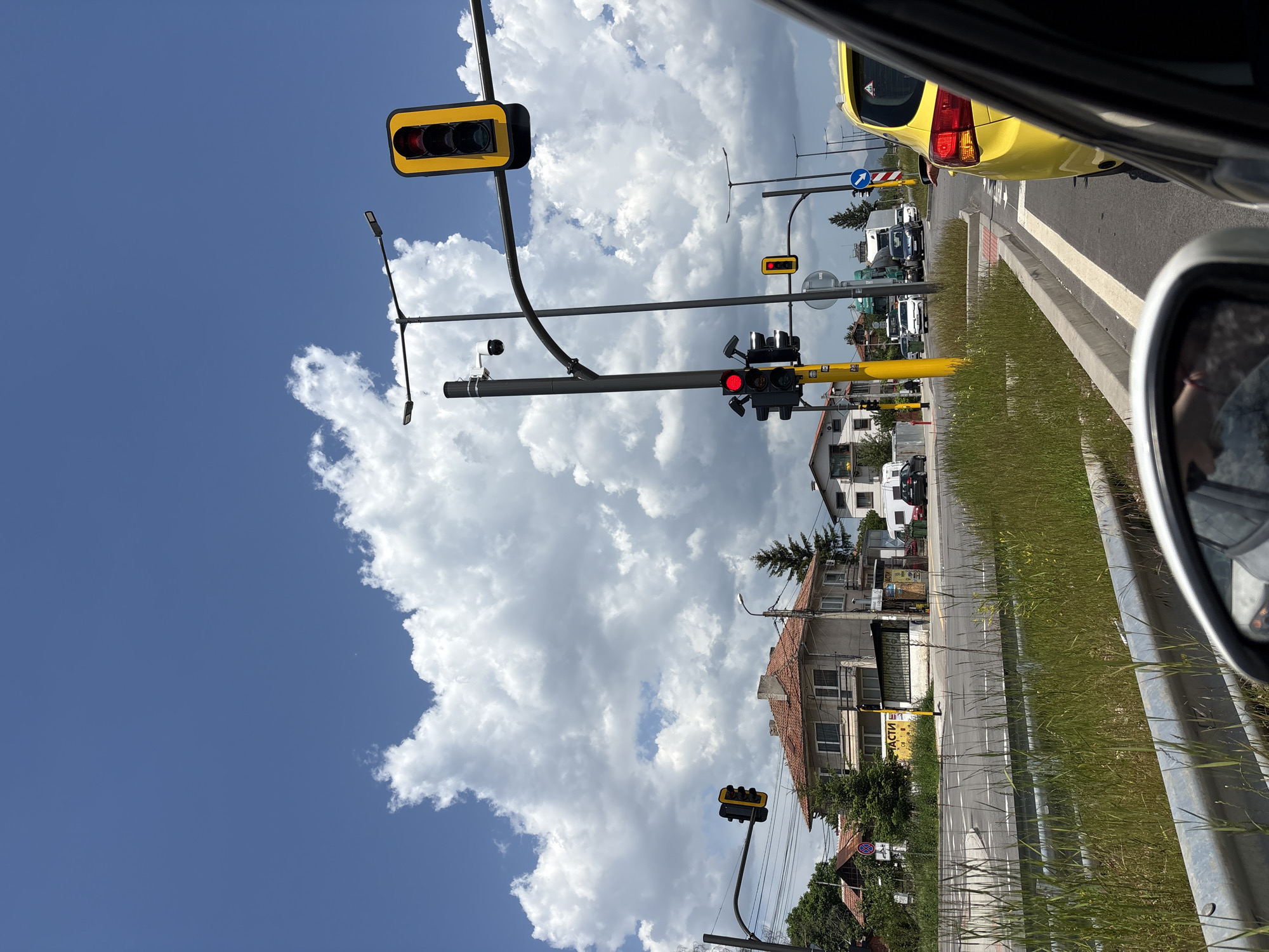 The image shows a view from inside a car, looking out onto a street scene in an urban environment. Multiple traffic lights are visible, all showing red lights. A yellow car is partially visible in the foreground to the right, and other cars and trucks can be seen further down the street. There are buildings visible on the right side of the street, and there is grass growing along the roadside. The sky is filled with cumulus clouds, contrasting with patches of blue sky.