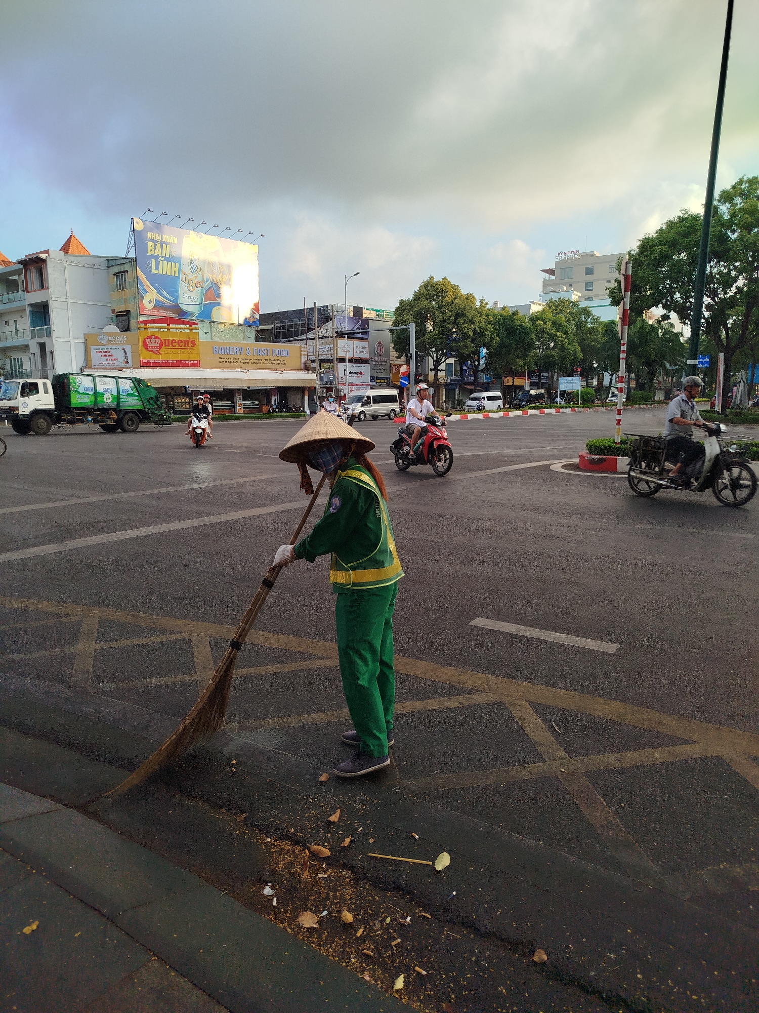 A street cleaner wearing a conical hat and green uniform is sweeping debris in a busy intersection in Ho Chi Minh City, Vietnam. Several motorbikes are traveling through the intersection, and a garbage truck is parked nearby. Buildings and billboards are visible in the background. The sky is overcast.