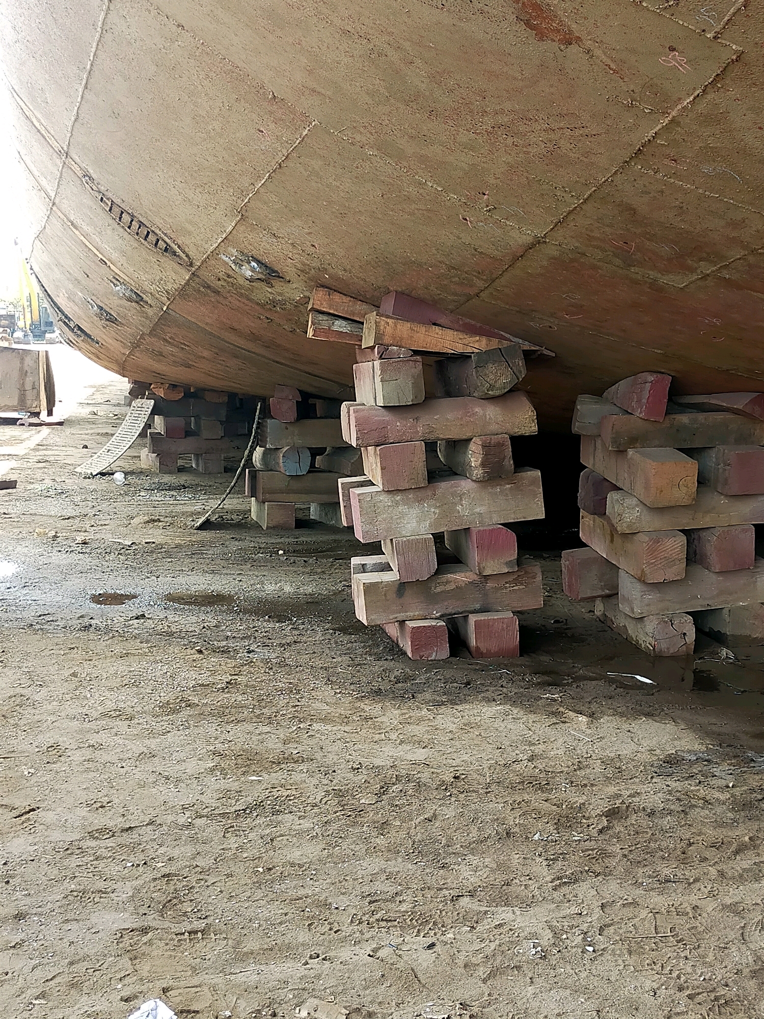 The image depicts the hull of a large ship resting on wooden blocks on a muddy, uneven surface. The ship's hull is a weathered, reddish-brown color, showing signs of corrosion and repairs. The wooden blocks are stacked in various configurations, providing support to the vessel. Some blocks are stained red, possibly from paint or other materials. In the background, construction equipment and a ladder are partially visible. The ground is covered in dirt, with several puddles of murky water scattered across it. The overall scene suggests a shipyard or dry dock environment in the city of Kamal, Indonesia.