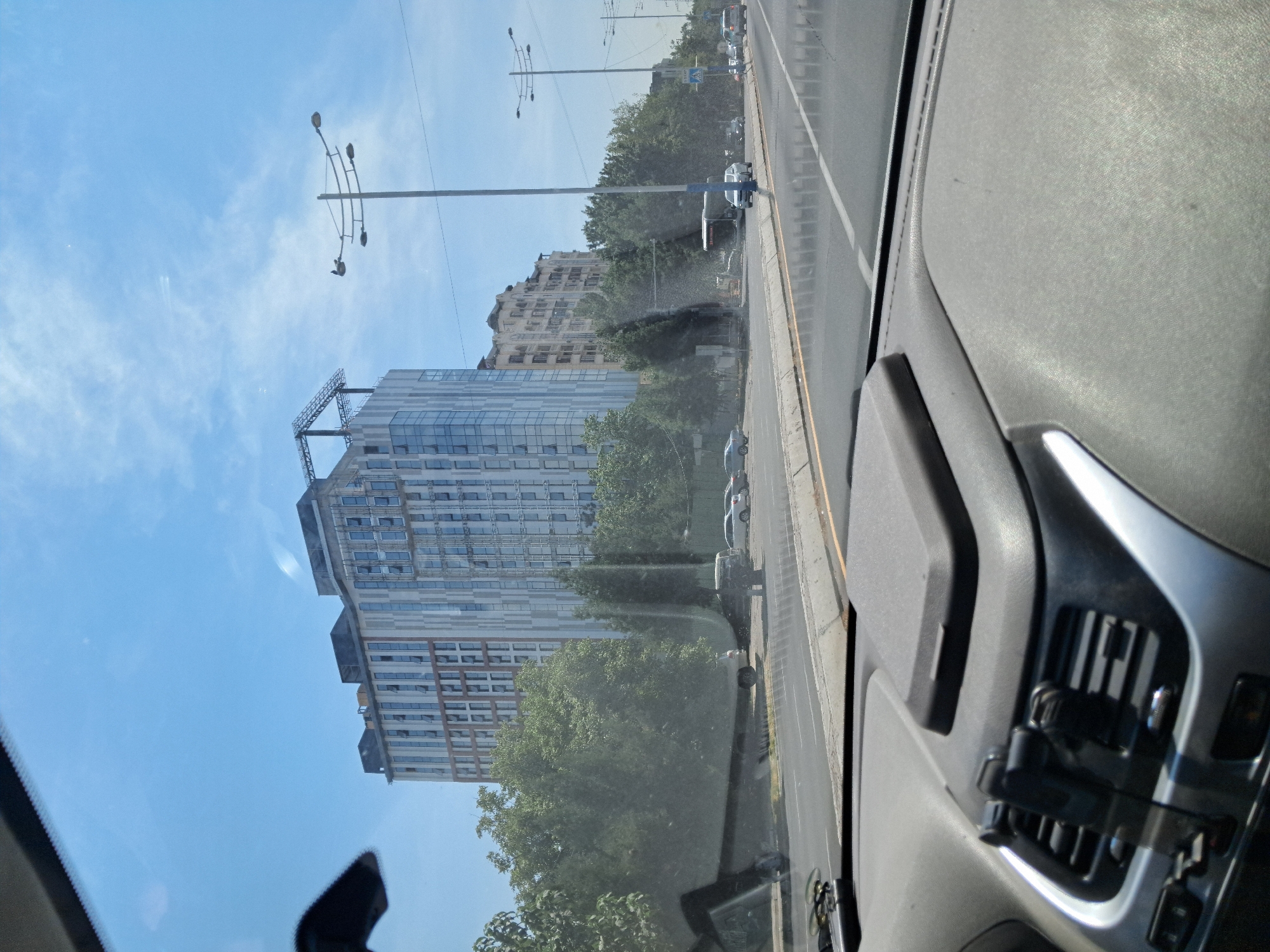 This is a photograph taken from inside a vehicle, looking forward through the windshield. The view encompasses a city street scene in Tashkent, Uzbekistan, with a modern, multi-story building dominating the background. The building features a facade of alternating light-colored vertical stripes and glass panels, with several windows visible. Above the building, a portion of a construction crane can be seen.
In the mid-ground, a line of trees separates the street from the buildings. Several cars are parked along the street, including a white van and several sedans. A bus is also visible on the road. The street itself is paved, with multiple lanes of traffic. Several streetlights with multiple lamps are positioned along the side of the road.
The foreground of the image shows the dashboard of the vehicle, with a black car mount and part of the air conditioning vent visible. The sky above is blue with scattered white clouds. The overall impression is a daytime scene in an urban environment.