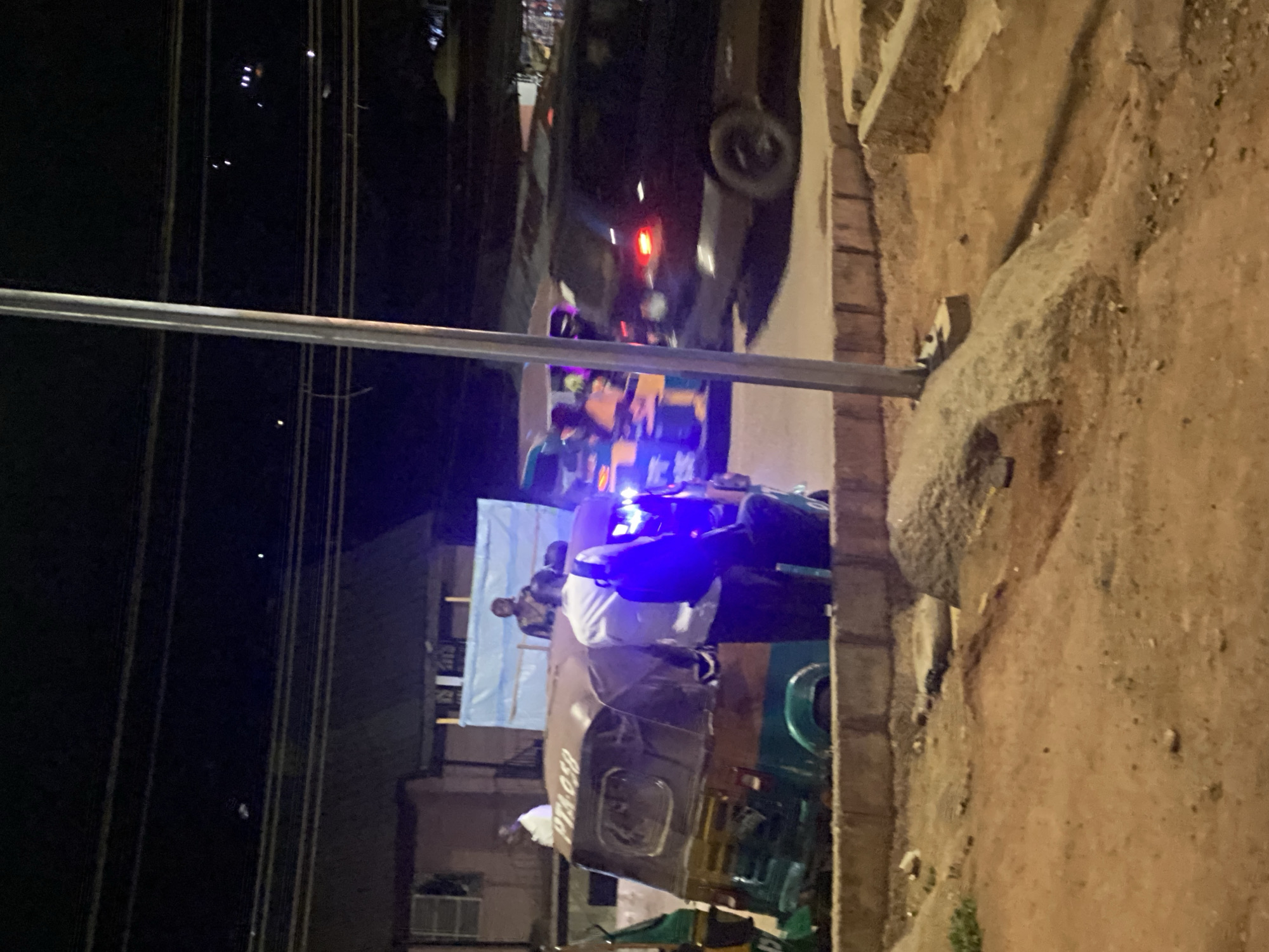 At night in what appears to be Gwong, Nigeria, the scene shows an area next to a road. There are a few people, their faces partially obscured, standing around and working on a vehicle, possibly a van or truck. The vehicle has a white cloth or sheet nearby, possibly to offer shade or for other purposes. There is also a car parked on the road. Wires of some sort are strung above, and the surroundings include buildings and ground with dirt and vegetation.
