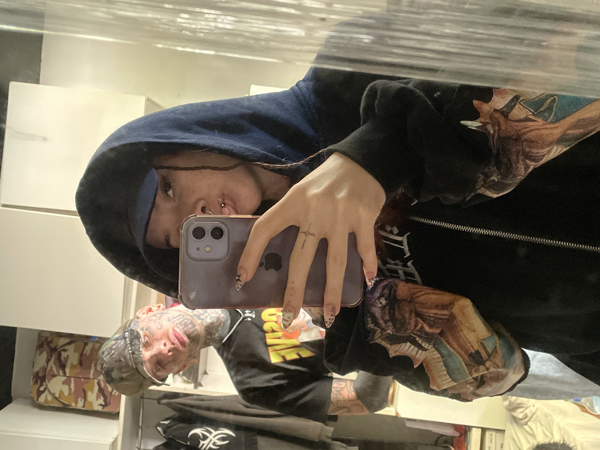 A person is captured in a mirror selfie, holding an iPhone with a light purple case. The main subject, positioned on the left side of the frame, wears a dark blue hoodie, with their face partially visible. They have dark hair, a septum piercing, and another small piercing on their nose, looking directly into the phone. Their hand, adorned with a small cross tattoo on the middle finger and long, pointed nails with black and white patterns, holds the phone. The sleeve of the hoodie features a large, colorful graphic design, possibly an abstract illustration or pattern.
Reflected in the mirror on the right side of the image is another person, possibly a male, with extensive facial tattoos covering his cheeks and neck. He wears a camouflage-patterned cap and a dark t-shirt with a yellow and orange graphic design on the front, which includes some illegible text. His arms also display numerous tattoos.
The setting appears to be indoors, likely a room or closet, indicated by white shelving and furniture visible in the background. A camouflage-patterned bag (pink, yellow, green hues) sits on a shelf behind the second person. The image is taken from a tilted perspective, characteristic of a casual mirror photograph. While the specific location isn't visually evident, the context provided states Buenos Aires, Argentina. Both individuals present a neutral to slightly serious expression.