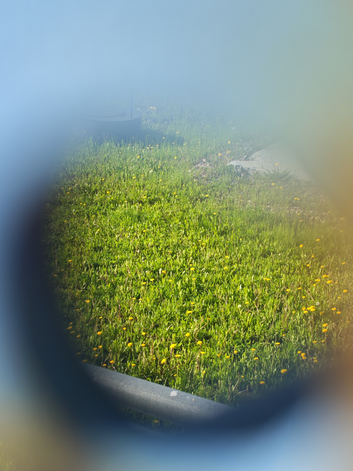 An overhead shot captures a patch of bright green grass dotted with numerous yellow dandelions. A portion of a gray metal bar is visible in the foreground, angled diagonally from the bottom left towards the center right. Beyond the grass, a light gray, flat surface is partially visible, possibly a concrete slab or a large stone. In the upper left portion of the image, a dark gray cylindrical object with a flat top emerges from the ground, with a thin, dark vertical pole extending from its center. The scene appears to be outdoors, with a soft, hazy blue sky filling the upper part of the frame, suggesting it is daytime. The image is framed by a blurred, dark, oval shape that encircles the central scene, hinting at a lens or viewpoint effect.