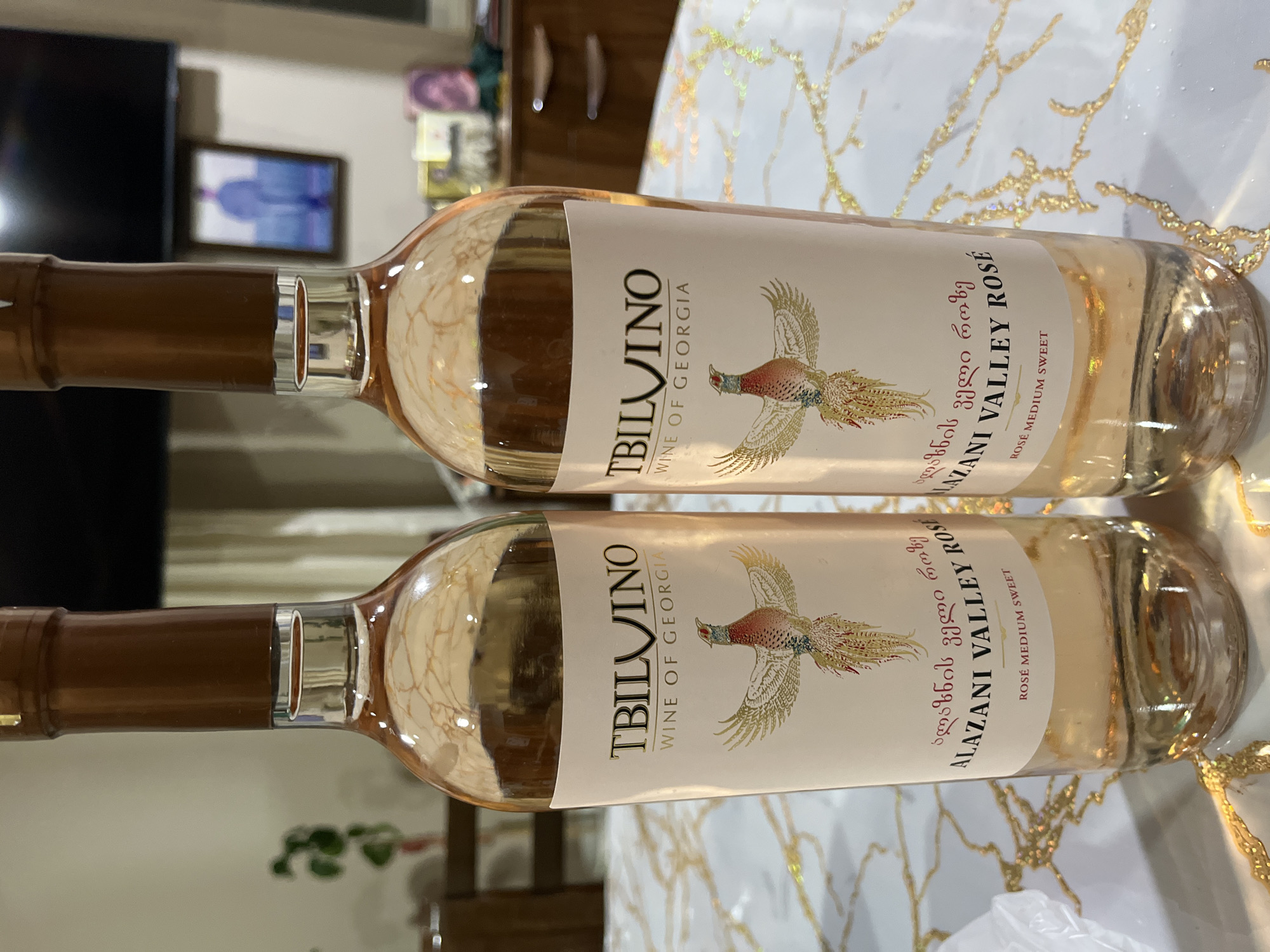 Two identical bottles of TBILVINO Alazani Valley Rosé wine, labeled "WINE OF GEORGIA" and "ROSÉ MEDIUM SWEET," are positioned side-by-side on a white surface with a prominent gold, swirling, patterned design. Each bottle is clear glass containing a pale rosé-colored liquid and sealed with a brown foil cap. The labels feature the brand name in black uppercase letters, Georgian script, and an ornate illustration of a pheasant with golden wings. The scene is an indoor setting in the city of Unknown, Unknown. In the blurred background to the left, a green potted plant and part of a wooden chair are visible. To the right, also blurred, a dark rectangular object resembling a television screen, a framed picture showing a person from behind, and a dark wooden cabinet with various indistinct items are present.