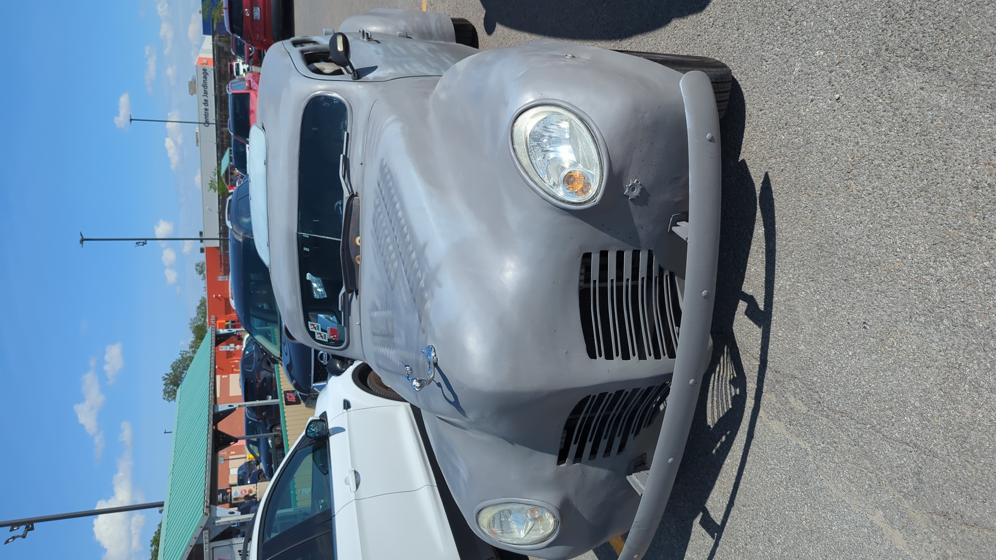 A vintage grey car, possibly undergoing restoration, is parked in a paved outdoor lot under a clear blue sky with scattered white clouds. The car features a distinctive chrome hood ornament and a slatted grille. In the background, several other vehicles are visible, along with the side of a building labeled "Centre de Jardinage" (Garden Center). The setting appears to be a commercial area in Laval, Canada.