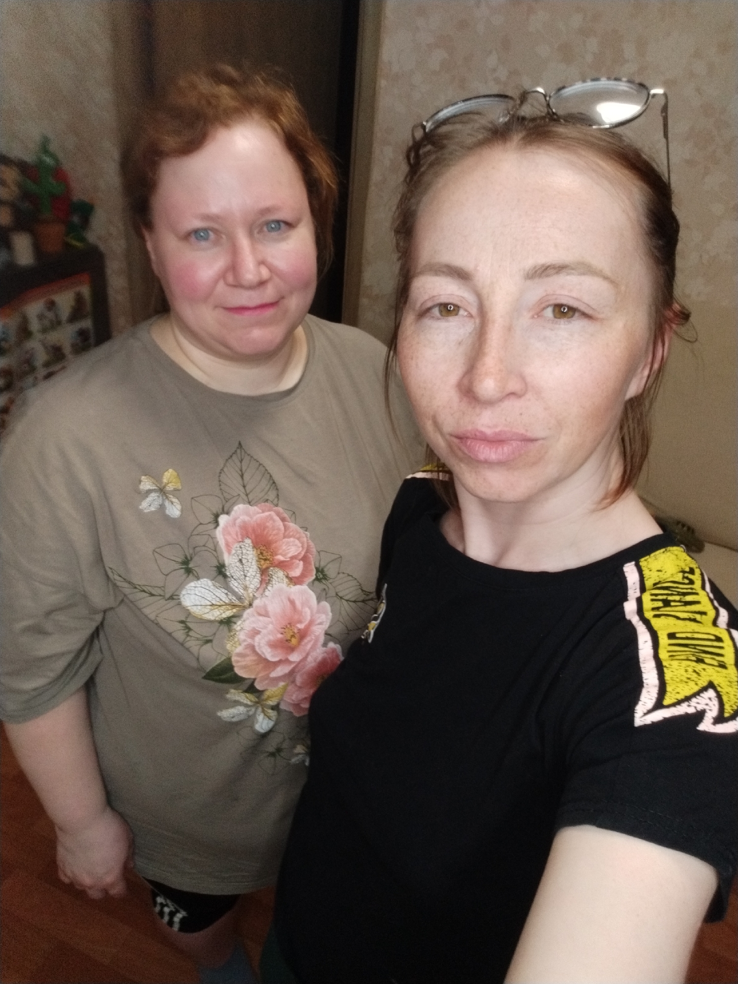 Two women stand close together indoors, taking a selfie. The woman on the left has fair skin, reddish-blonde hair, and blue eyes. She is wearing a light brown t-shirt with a floral design on the front. The woman on the right has fair skin with freckles, brown eyes, and reddish-brown hair pulled back. She has glasses resting on her head and is wearing a black t-shirt with a yellow and white design on the shoulder. They are both looking towards the camera.
Behind them, the background appears to be a simple interior with light-colored walls. To the left, a piece of furniture with various decorative items is visible. The location is Chaikovsky, Russia. The lighting is soft and diffuse, typical of indoor lighting.