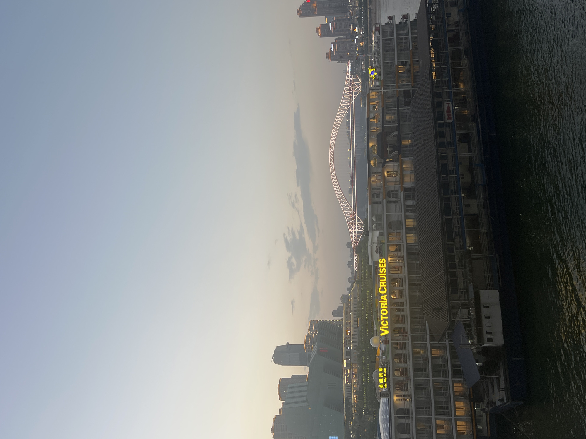 A wide shot captures a twilight scene in Yuzhong District, China, featuring a multi-decked cruise ship docked on a dark body of water. The cruise ship prominently displays illuminated yellow text reading "VICTORIA CRUISES" and "美維凱迪". Behind the ship, a modern cityscape rises, characterized by numerous tall buildings with illuminated windows. A distinctive arched bridge, outlined with white lights, spans across the background, connecting different parts of the city. The sky is a gradient of soft blues and grays, with a few wispy clouds. The overall impression is one of urban development and riverine transportation at dusk.