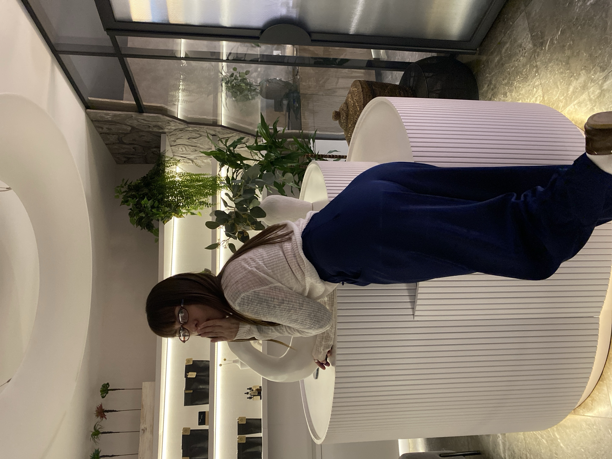 An indoor scene in Votkinsk, Russia, displays a woman interacting with products in a modern retail or showroom environment. The image, originally rotated, shows a woman with long dark hair and glasses, dressed in a light-colored, possibly off-white, long-sleeved top or cardigan and dark blue, wide-leg pants. She is standing and leaning over a white, horizontally ribbed display counter, with her hand partially covering her mouth, suggesting contemplation, curiosity, or a subtle reaction.
The setting is characterized by a clean, contemporary design, predominantly featuring white furniture and decor. Prominent elements include multiple white, cylindrical or curved display pedestals and counters with distinct horizontal ridging. On one such counter, a white, swan-shaped decorative object is visible. The space incorporates natural accents, such as various artificial plants and greenery, including ferns, arranged in different areas.
Behind the woman, shelves are illuminated with strip lighting, showcasing small, dark-colored items (possibly product packaging or samples) alongside some lighter ones. Below these, small artificial palm-like plants are arranged on a lower shelf. The background features a glass and dark metal framework, resembling a ceiling or high partition, and a section of wall with a textured, stone-like finish. A woven basket is also visible in the background. The flooring appears to be a light-colored, reflective surface, possibly polished stone or tile.
The artificial lighting suggests an indoor setting where the time of day is not a key factor, creating a sophisticated and well-lit ambiance. While there are small labels on the displayed items, no specific text is clearly legible to identify products or branding. The overall scene depicts a quiet, engaging interaction within a stylish commercial space.